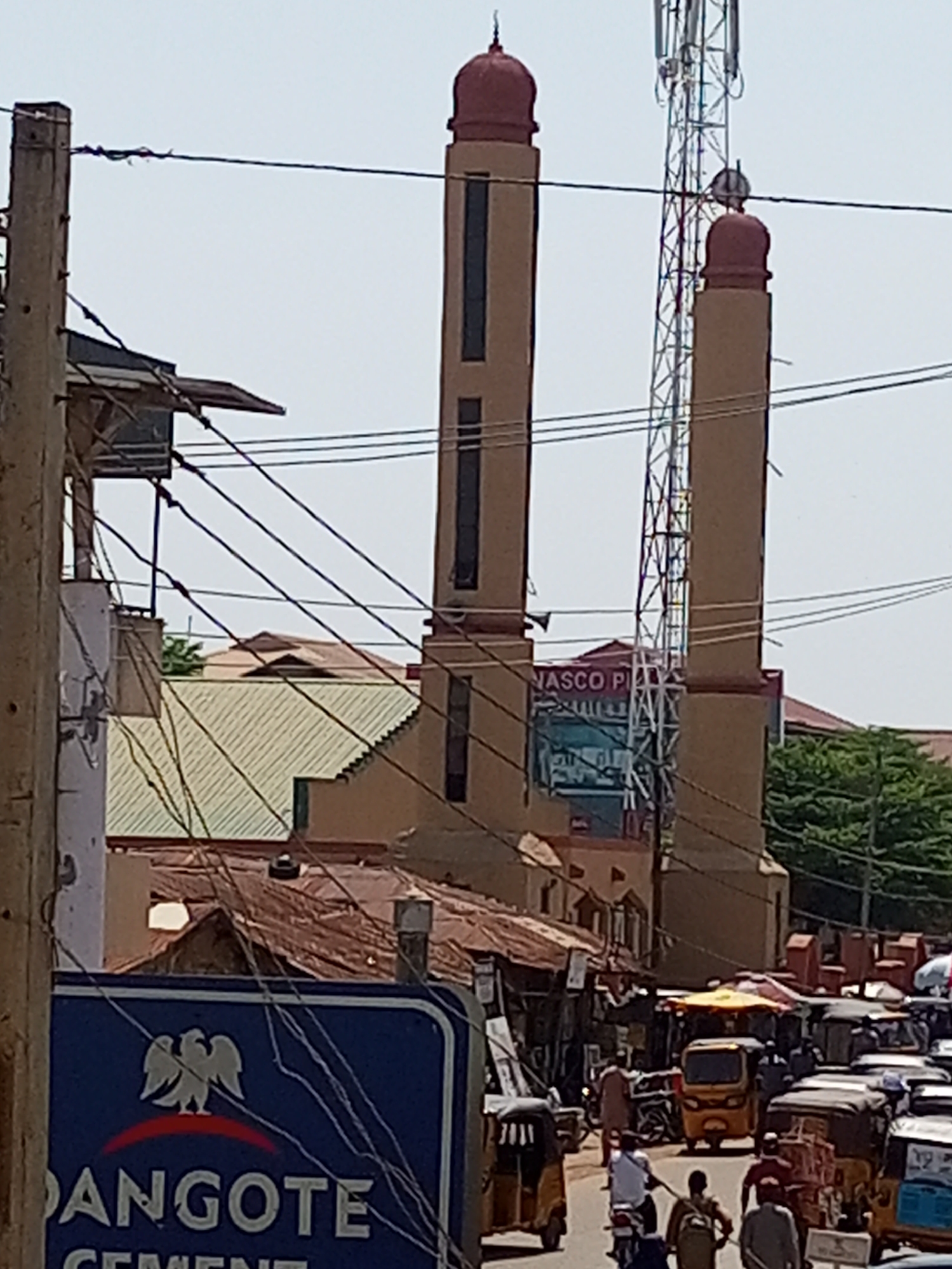 A busy street scene in Maiburiji, Nigeria, is captured during daylight hours under a bright, clear sky. In the background, two prominent minaret-like structures with reddish-brown domed tops stand tall, flanking a slender metal telecommunications tower. Numerous tangled electrical wires crisscross the sky and connect to wooden utility poles that line the street. Below the minarets, a building with a light-colored corrugated roof is visible, and a commercial sign bearing the partially visible text "NASCO PI" can be seen between the minarets.
The foreground features a bustling street filled with people, motorcycles, and yellow auto-rickshaws (locally known as Keke Napep), indicating an active urban environment. Various buildings with different roof types, including corrugated metal, and market-like stalls line the roadside. A large blue sign in the lower left foreground prominently displays a white eagle logo and the partially visible text "DANGOTE CEMENT." Green foliage from trees is visible in the background, adding a touch of nature to the otherwise built environment.