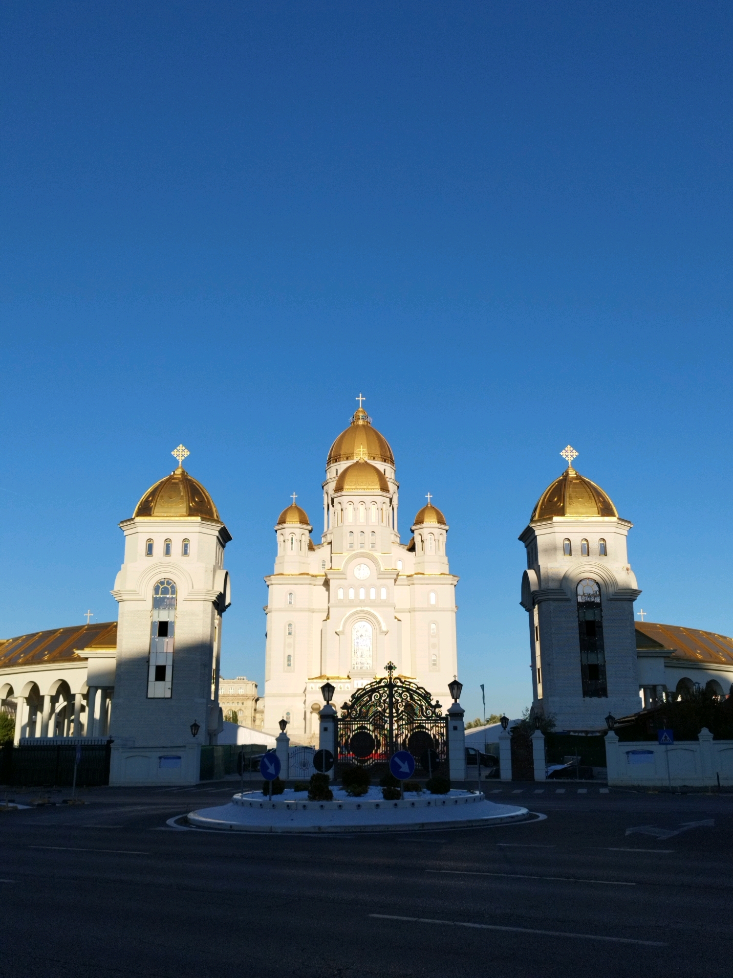 The image shows the Patriarchal Cathedral of Bucharest, Romania, from a low-angle view in daylight. The cathedral, a prominent religious landmark, is characterized by its white facade and multiple golden domes topped with crosses. The central dome is the largest, flanked by two smaller domes and two bell towers. The foreground features a street intersection with a roundabout, a decorative iron gate, and traffic signs. The sky is a clear, vibrant blue. The scene is devoid of people and shows no signs of activity beyond the static architectural elements and road infrastructure.