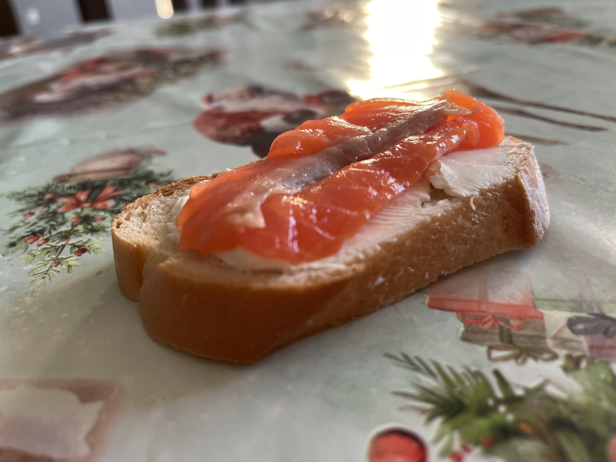 A close-up shot shows a canapé on a tablecloth. The canapé consists of a slice of bread topped with white cream cheese and several slices of smoked salmon, including a darker strip in the middle. The tablecloth has a light green base with a pattern of Christmas-themed decorations like ornaments, gift boxes, and pine branches. The lighting is bright, with sunlight reflecting off the salmon. The location in the city of Chaikovsky, Russia is unknown.