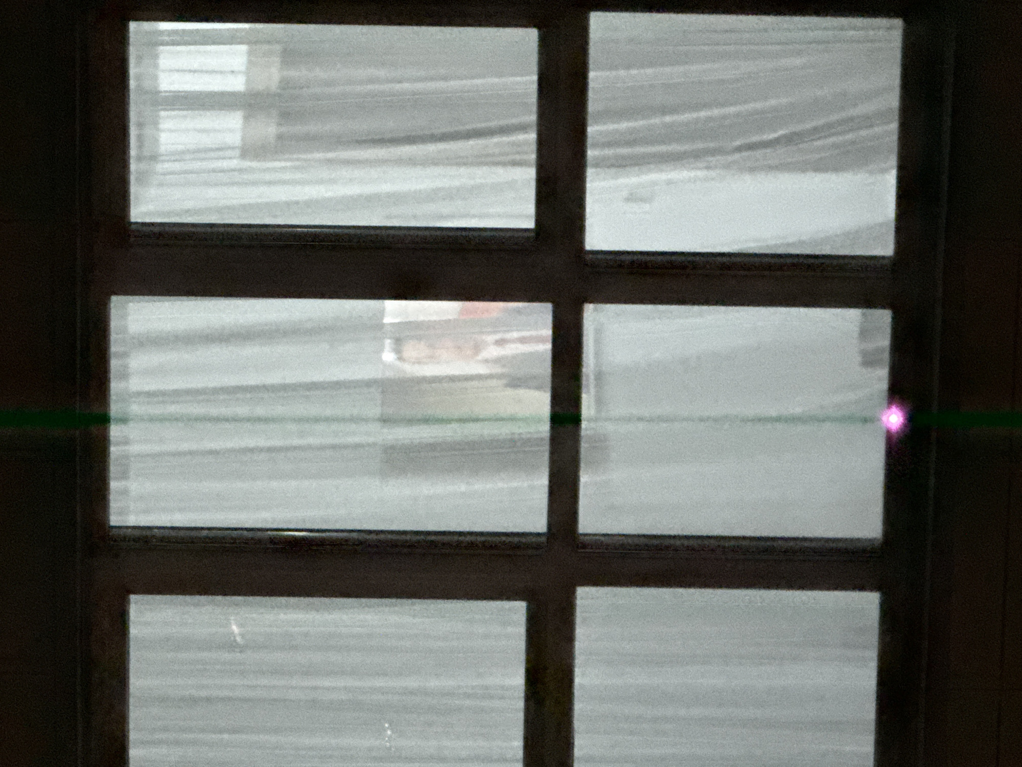The image shows a window divided into six panes, through which a snowstorm is visible. The snow appears to be heavy, with horizontal streaks indicating its movement. Outside, the scene is obscured by the blizzard, but a few indistinct shapes suggest buildings or structures. A green laser beam with a bright pink dot is visible horizontally across the window. The specific location in Khimki, Russia, cannot be determined from the image alone.