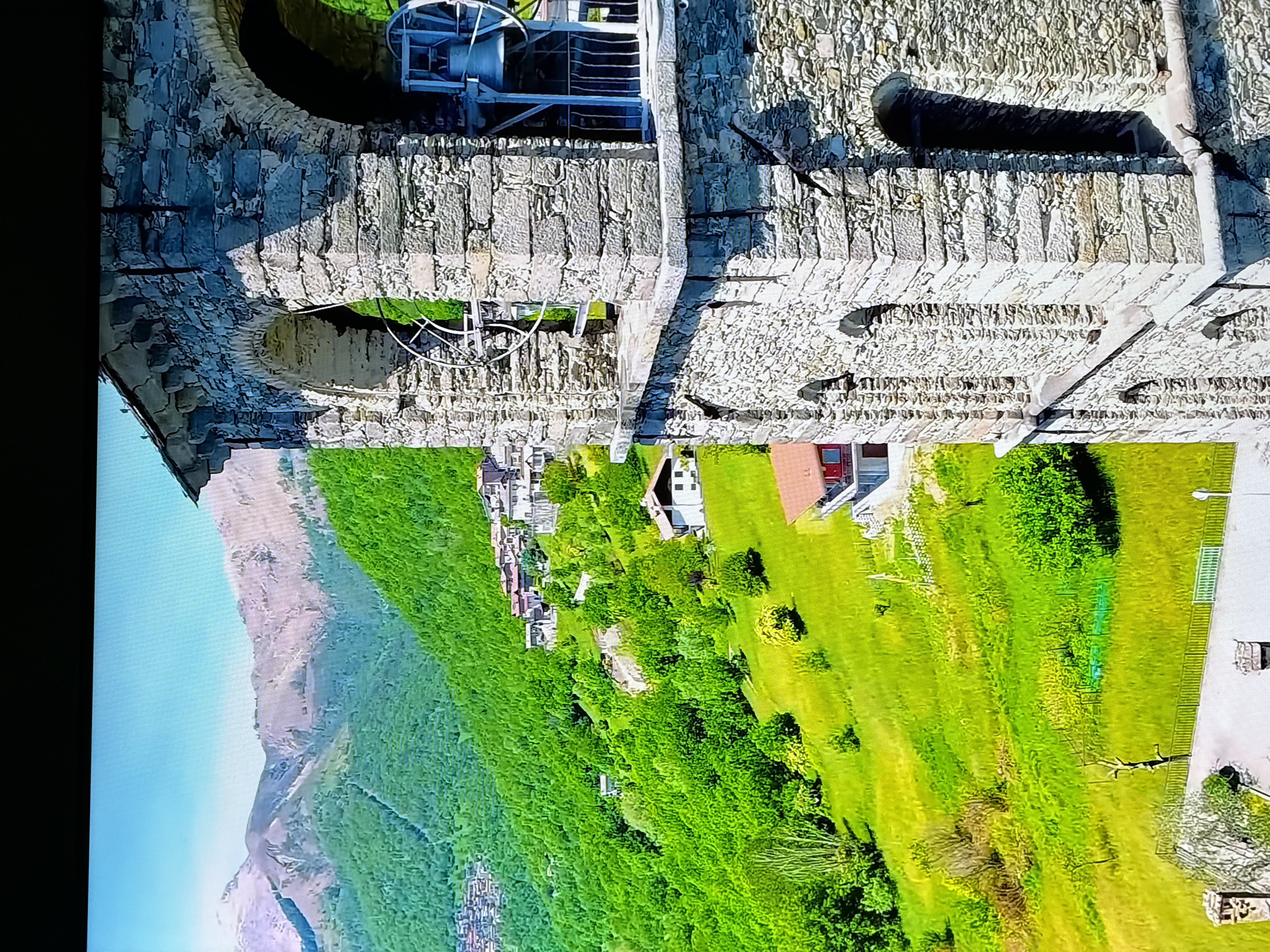 The image shows a stone bell tower with arched openings overlooking a lush green mountainous landscape. The tower itself is made of rough-hewn stone blocks and features an ornate ledge. Through the arched openings, a large bell and its supporting structure are visible. In the background, a village or town is nestled amongst the densely forested hills, with red-roofed houses and buildings scattered across the slopes. Beyond the hills, majestic mountains with snow-capped peaks rise against a clear blue sky. The foreground shows a portion of the tower and the verdant, undulating terrain leading up to it. The scene appears to be a picturesque view of a historic structure in a rural, mountainous setting. There are no people visible in the image. This scene is not in Chirchik, Uzbekistan.