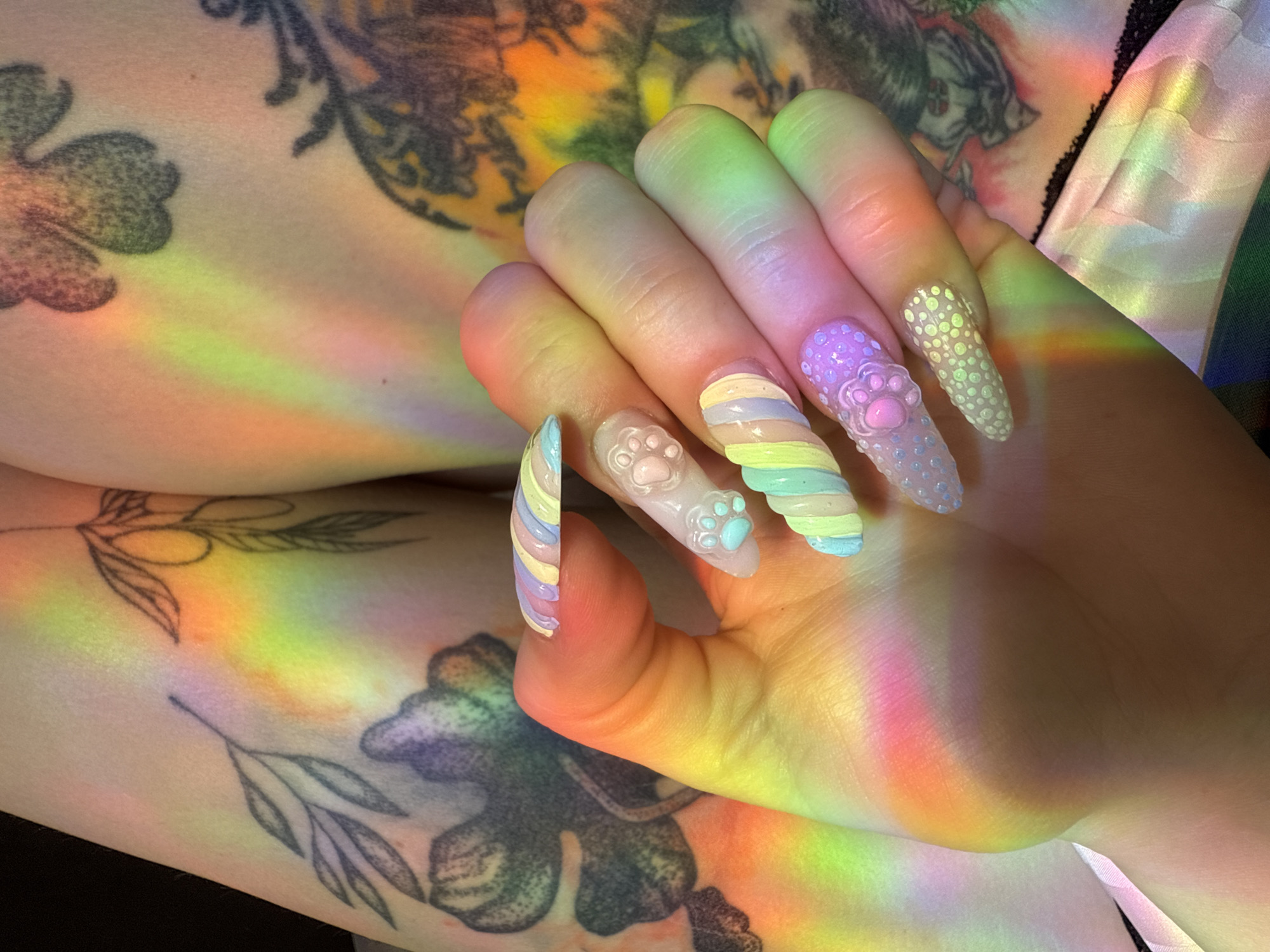 A close-up image shows a person's forearm and hand adorned with tattoos and elaborately decorated nails. The arm, resting on an indiscernible surface, features a collection of tattoos depicting floral and other designs. The nails are elongated and styled with pastel colors, stripes, polka dots, and 3D paw print embellishments. A prism effect casts rainbow-colored light bands across the skin, nails, and tattoos. The location is Unknown, Unknown.