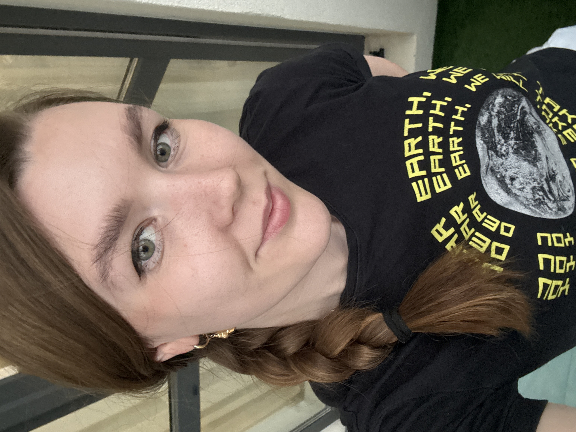A woman with fair skin and light brown hair styled in a braid is looking at the camera. She has green eyes and is wearing a black t-shirt with yellow text and a graphic of the earth on it. The background appears to be an indoor setting, with a window frame visible behind her head. There is also a glimpse of green foliage in the background. There is no indication that the picture was taken in Izhevsk, Russia.