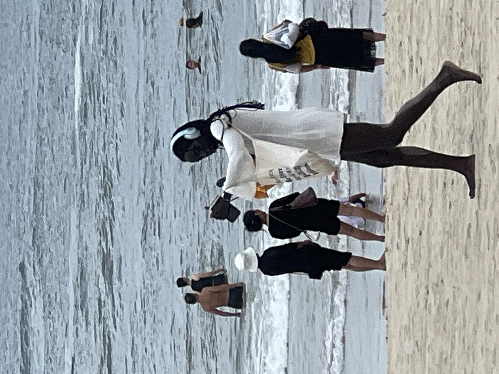 The image shows a beach scene at the ocean in Đà Nẵng, Vietnam. In the foreground, a person with dark skin, wearing a white shirt and blue headphones, walks on the sand carrying a white bag. Behind them, two women are standing in shallow water, one wearing a white hat and black clothing, the other in black. Further out in the water, several people are visible, some swimming or wading. The water is choppy with white waves. The beach sand is light brown.
