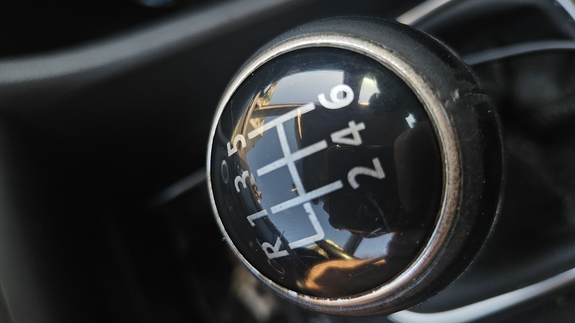 The image shows a close-up view of a black manual car gear shifter with white markings for the gear positions. The gear pattern is visible, indicating a 6-speed manual transmission with a reverse gear. The numbers 1, 2, 3, 4, 5, 6, and R are clearly marked, arranged in a standard H-pattern. Reflections are visible on the glossy surface of the gear knob, showing parts of the car's interior, including what appears to be a dashboard and a person's hand. The background is dark and out of focus, suggesting the interior of a vehicle. The lighting suggests it could be daytime, given the reflections, but the overall scene is dimly lit. The image focuses solely on the gear shifter, highlighting its design and function within a car. No specific location cues are present, and no weather conditions are discernible. The image does not capture any people directly, only reflections of a hand.