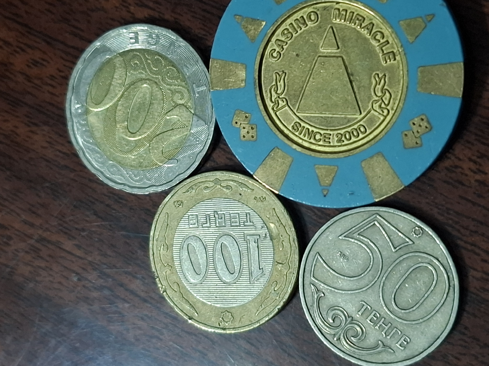 The image shows four casino tokens and coins placed on a wooden surface. The objects are in the center of the frame, and the background is a dark, grainy wooden table. Three of the objects appear to be coins, two of which have the number "100" and "50" prominently displayed. The third coin has a circular design with what looks like wavy lines. The fourth object is a blue and gold casino token with dice symbols and text that reads "CASINO MIRACLE SINCE 2000". There are no people or actions depicted in the image. The lighting suggests an indoor setting with the light source coming from above and slightly to the left. The location in Almaty, Kazakhstan, is not discernible from the image itself.