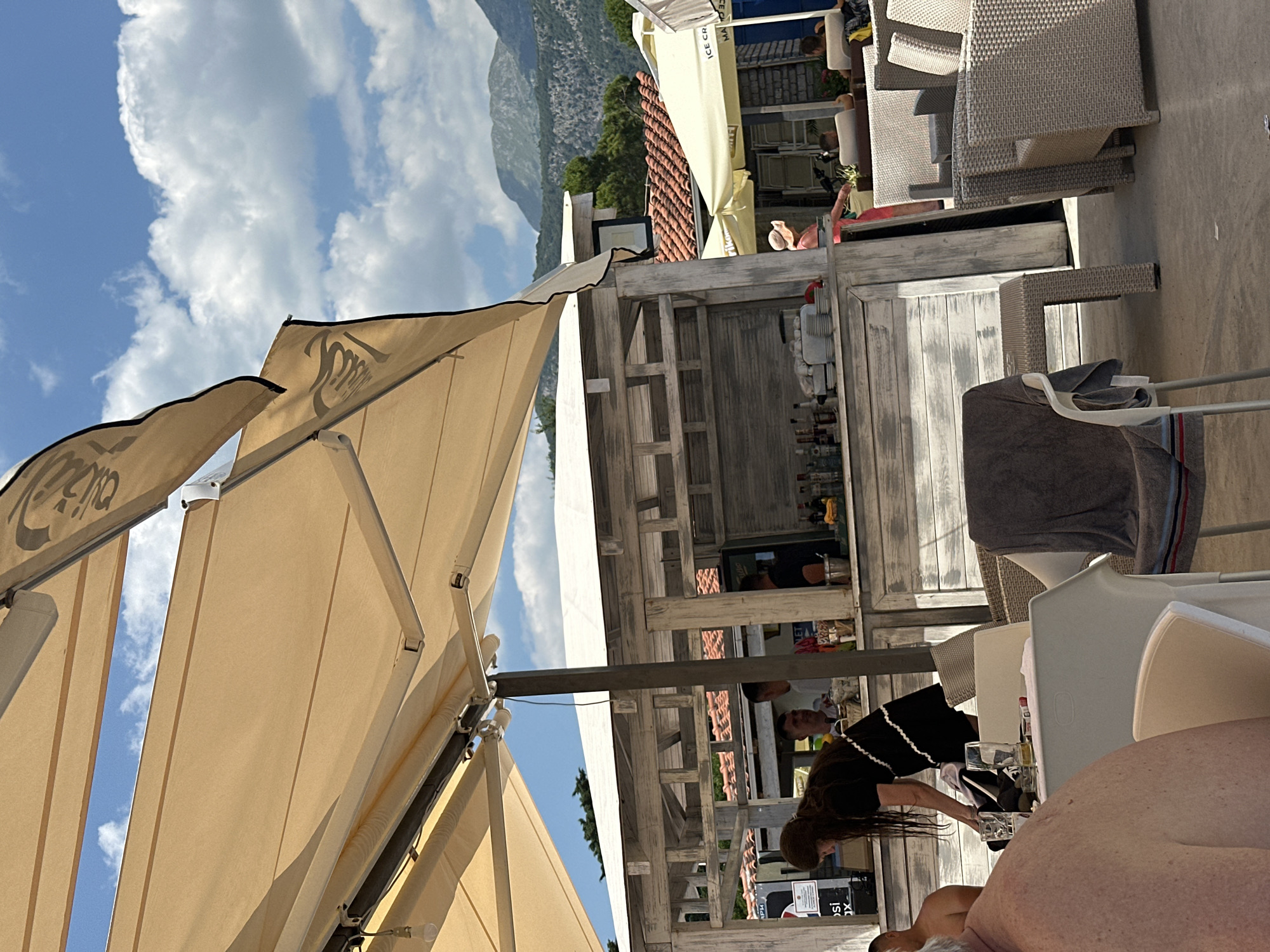A group of people are seen at an outdoor cafe in Budva, Montenegro, on a sunny day. The cafe features white wooden structures and beige awnings providing shade. In the foreground, a person with long dark hair wearing a black dress is visible from the side, interacting with items on a table. Several tables with white chairs and wicker seating are arranged around the cafe. In the background, a bar area is set up with bottles and glassware. Other patrons are seated at tables or standing near the bar. Mountains and lush greenery form the backdrop, with a bright blue sky and scattered clouds overhead. The scene appears to be a lively and relaxed atmosphere at a beachfront establishment.