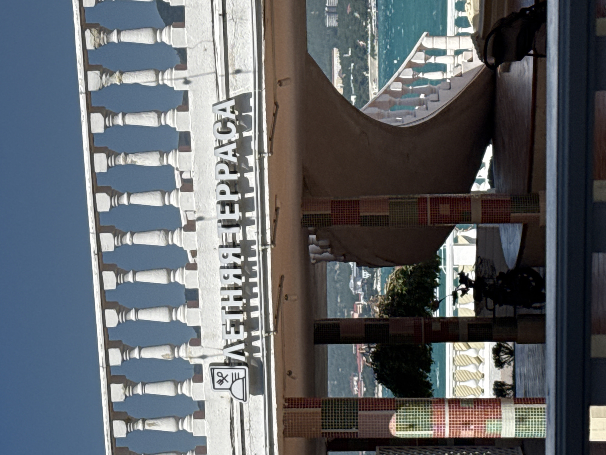 The image shows a sign with the Russian text "ЛЕТНЯЯ ТЕРРАСА" (Summer Terrace) above an entrance to a venue. To the left of the text is an icon representing a restaurant. The sign is mounted on a white architectural structure with a balustrade above it. Below the sign, a spiral staircase is visible, leading down to an outdoor area. The background reveals a body of water, likely the Black Sea, with buildings and trees on a distant hillside, indicative of Gelendzhik, Russia. The scene suggests a waterfront establishment offering outdoor seating.