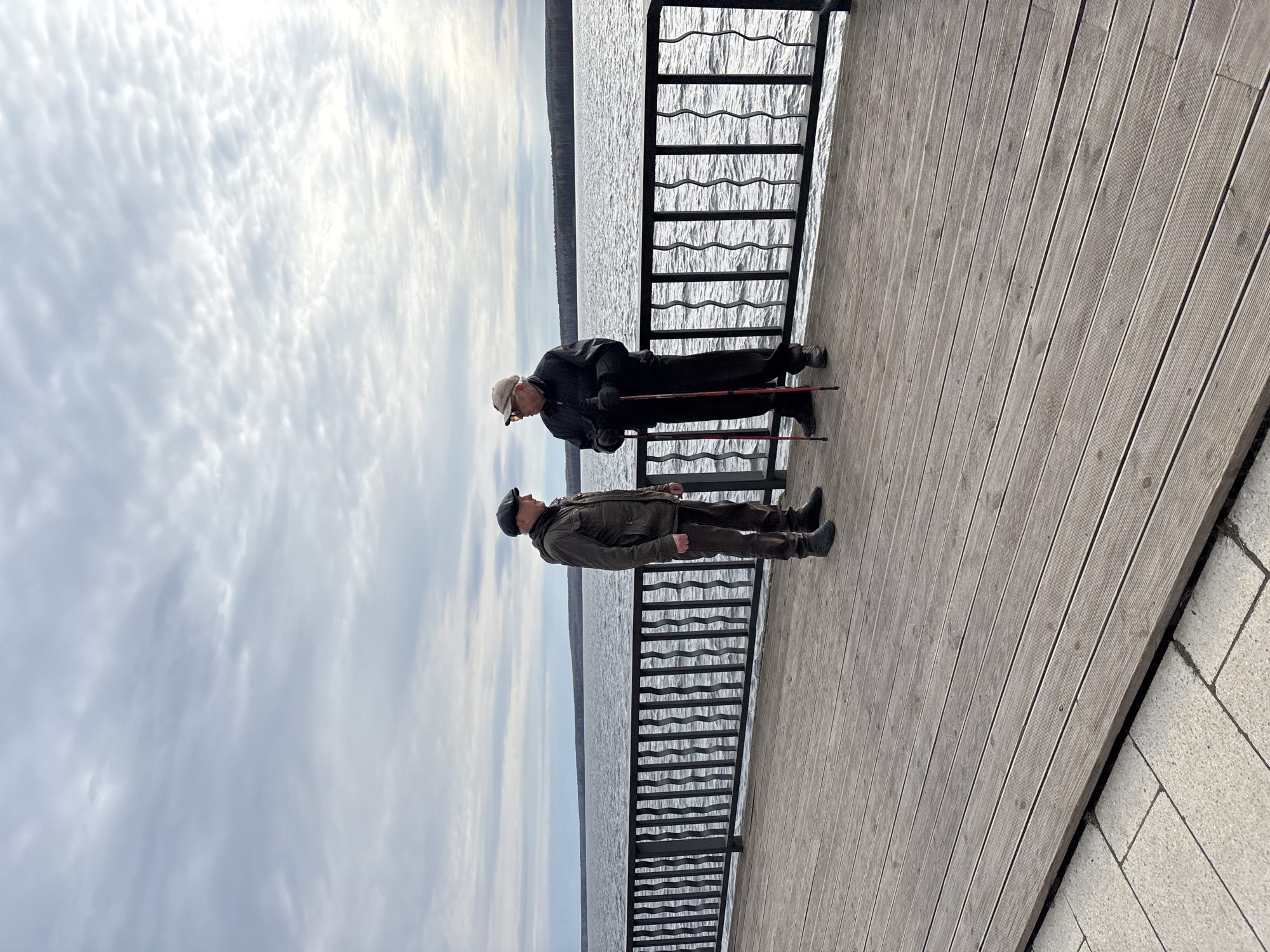 Two men are standing on a wooden pier with a black metal railing, facing each other and talking. The pier is located on the shore of a large body of water, likely the Votkinsk Reservoir, in Chaikovsky, Russia. Overcast skies are visible above the water. One man is wearing a dark jacket, light-colored cap, and sunglasses. He is holding what appears to be a walking stick. The other man is wearing a dark cap and a brown jacket. The pier is made of light-colored wooden planks and is connected to a paved area on the right side of the frame.
