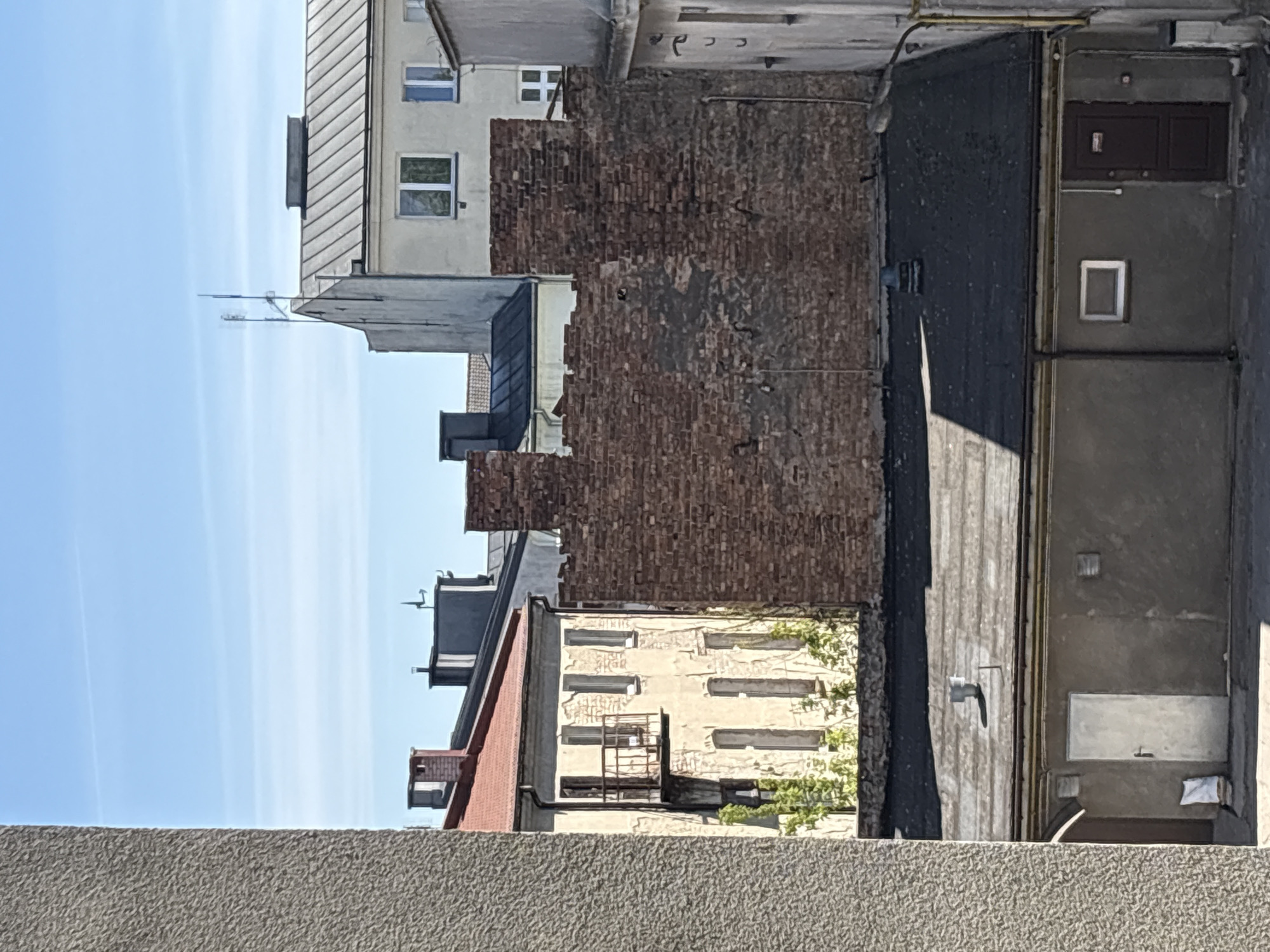 The image depicts a view of urban architecture in Mława, Poland. A concrete wall is visible in the foreground. Behind it are a series of buildings of varying heights and architectural styles. One building features a brick wall with visible damage. Another building has rows of windows. A metal ladder is seen on one of the buildings. There is also a rooftop with a red-tiled section. A blue sky is present in the background.