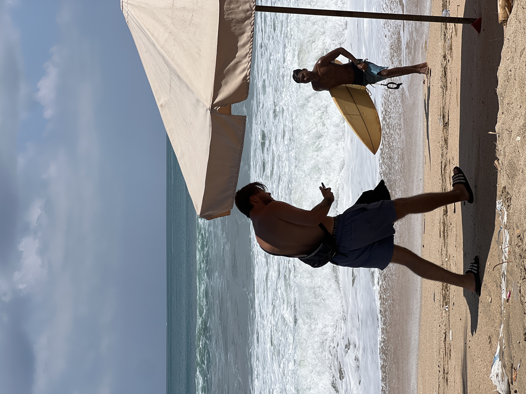 On a beach in Kuta, Indonesia, two men are visible. The man in the foreground is walking left to right, looking down at his phone. He is wearing blue shorts and a black fanny pack. He is also wearing black slide sandals. Behind him, a second man is walking right to left, carrying a yellow surfboard. He is wearing blue swim trunks and appears to be looking towards the first man. The ocean in the background has moderate waves, and there is a large beach umbrella on the right side of the frame. The sand appears to have some litter.