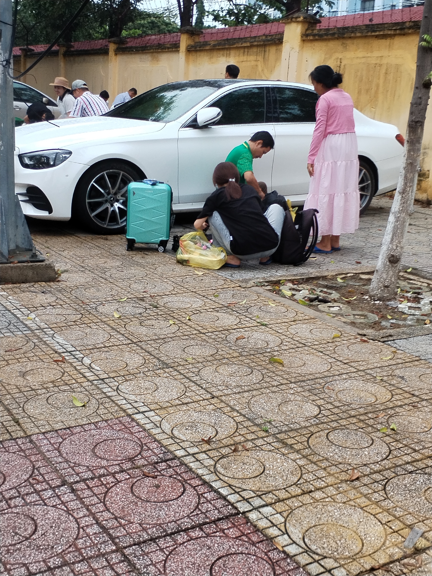 The image depicts an outdoor urban scene, likely in Nha Trang Ward, Vietnam, centered around a white sedan car parked on a patterned tiled sidewalk.
A group of people is gathered near the car. In the foreground, two individuals are crouched down. One person, seen from behind in a black top and light pants, is looking into a yellow plastic bag. Adjacent to them, a person in a green t-shirt is also bent over near the car. A light blue, patterned carry-on suitcase on wheels is positioned between them and the car, and a black backpack lies on the ground nearby. Standing to the right, a woman in a long, layered pink dress and a matching pink long-sleeved top observes the crouched individuals. Further back and partially obscured by the car, other people are visible, including a man in a plaid shirt and a hat, and a woman wearing a straw hat.
The primary activity appears to be packing or unpacking items into or from the car, consistent with travel, indicated by the luggage. The setting includes a distinctive sidewalk with circular and grid patterns in earthy tones. Behind the car, a yellow-painted wall with a red coping and barbed wire on top forms the backdrop. A tree trunk stands on the right side of the frame, and a grey utility pole is on the far left.
The scene takes place during the daytime, under what appears to be diffused or overcast lighting conditions. The ground is dry. No visible text is discernible in the image.