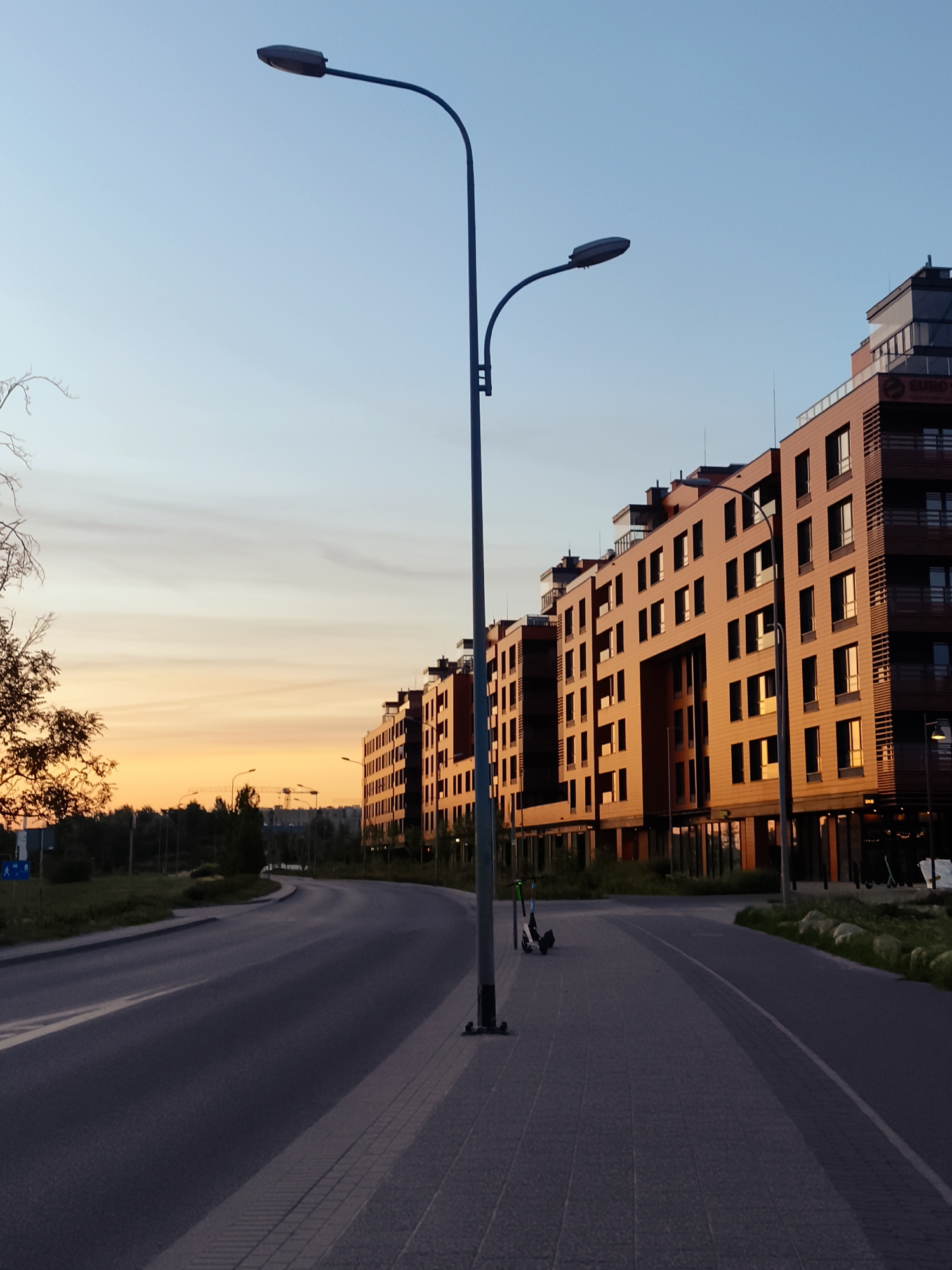 This is a medium-angle, eye-level, outdoor, vertical, color photograph taken at dusk. The scene depicts a modern urban environment with a focus on architecture and infrastructure.
In the foreground, a paved sidewalk runs along the right side of the image, bordered by a street on the left. The street curves gently into the distance. Two electric scooters are parked on the sidewalk near the center of the frame. A tall streetlamp with a modern design is positioned prominently on the sidewalk.
Dominating the right side of the image are several contiguous multi-story buildings with contemporary architectural features. The buildings are clad in warm-toned material, possibly wood or brick, and feature numerous windows, some of which reflect the sunset. The architecture suggests a residential or commercial complex. The "EURO SP. Z O.O." logo is visible on one of the buildings.
The sky transitions from a clear, light blue at the top to a vibrant orange and yellow hues near the horizon, indicative of a sunset. Silhouetted against the sunset are trees and distant structures, including what appear to be construction cranes, suggesting ongoing development in the area. A single tree with bare branches is visible on the far left, partially framing the sky.
The overall impression is one of a modern, developing urban area at the end of the day, with the warm tones of the sunset contrasting with the cool tones of the sky and street. The location is identified as the area around the Euro Sp. z o.o. building in Gdańsk, Poland.