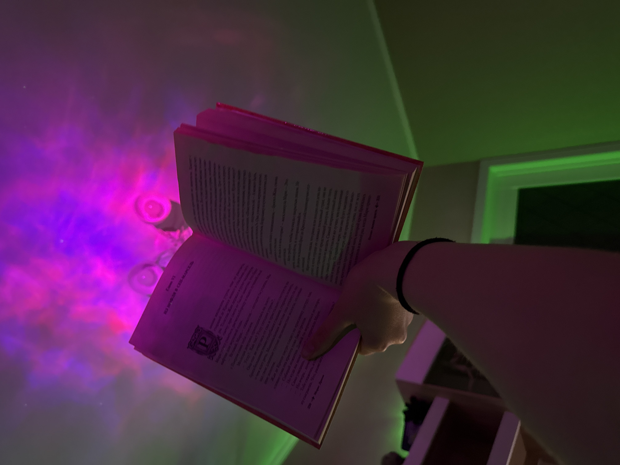 A person is holding an open book in front of them. The book is written in Russian and is being illuminated by what appear to be colorful, light projections. The room has a green and pink hue. A shelf is visible in the background. The content of the book seems to be a chapter titled "Camme 93. Магирля в сеM-МАРСЕЛЬ" (Mangirlya in Saint-Marcel) implying the story's potential location.