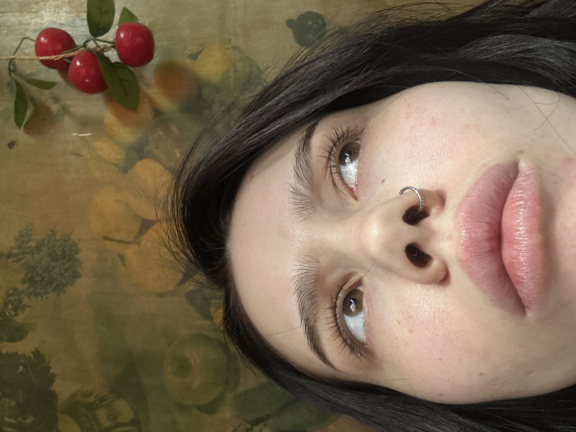 This image depicts a close-up, low-angle shot of a young female subject. Her face is partially visible, from below the nostrils to above the forehead, with dark hair framing her head. She possesses brown eyes, which are directed upwards and towards the right side of the frame. Her eyebrows are dark and prominent. A silver-colored hoop nose ring, adorned with small stones, is present in her right nostril. Her lips are visible, appearing full and slightly parted, with a mild sheen. The subject's skin exhibits minor textural variations consistent with pores or freckles.
The background consists of a patterned surface, possibly wallpaper or textile, in faded shades of green and yellow-orange, featuring abstract fruit-like shapes and foliage motifs. In the upper right quadrant, a decorative item resembling a cluster of three artificial red cherries with green leaves, suspended by a thin, light-colored string, is visible against the background. The scene is located in Aktanysh, Russia.