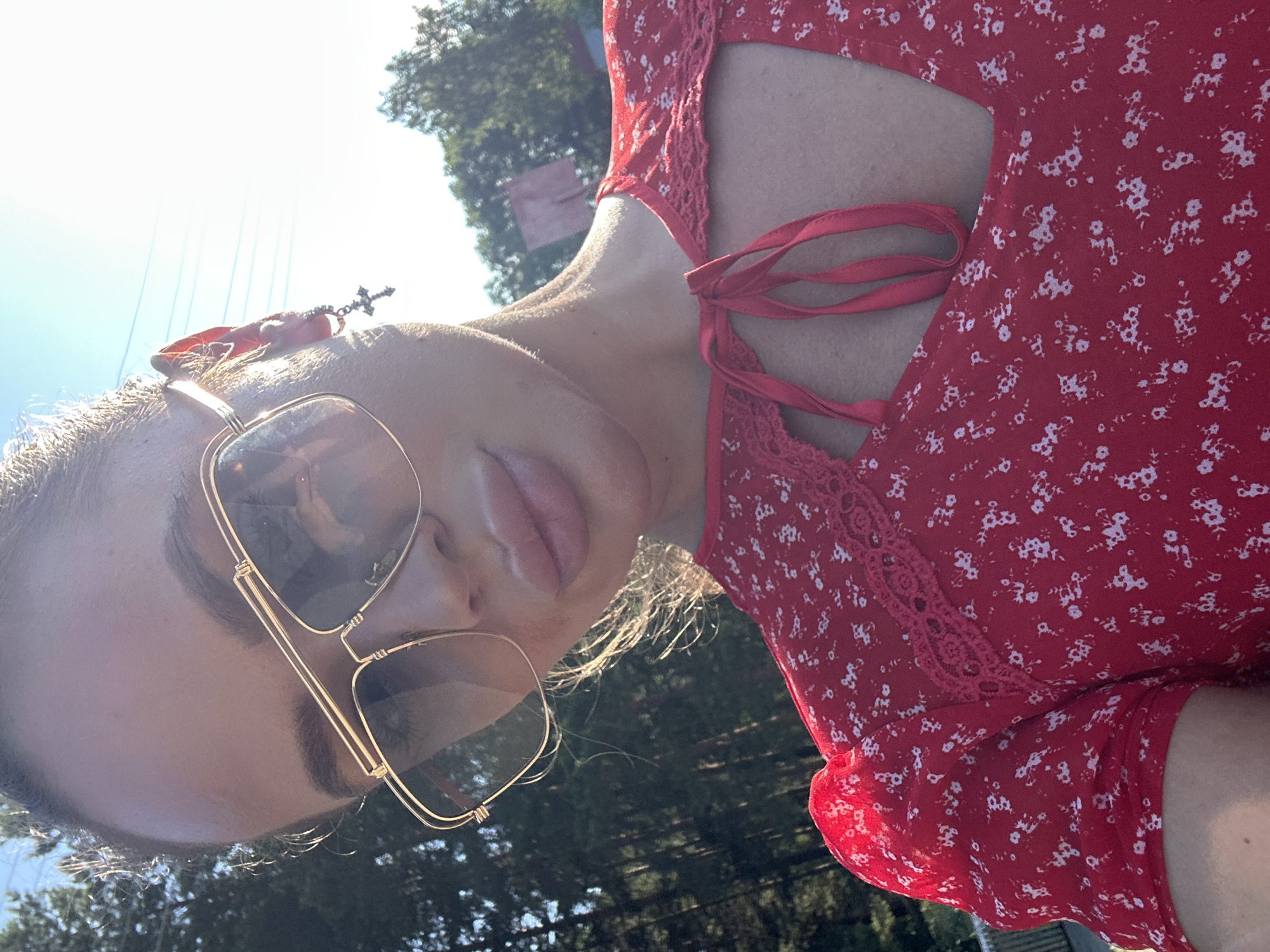 An outdoor, close-up, waist-up shot shows a young woman with her head turned to the right. She is wearing large, gold-rimmed aviator sunglasses and a red floral print dress with lace trim. The woman has a red ribbon tied in a bow at the neckline of her dress. Her hair is pulled back. A cross earring hangs from her right ear. Sunlight casts shadows on her face and illuminates her hair. The background shows trees and a bright sky. The image was captured in Duvankul', Russia.