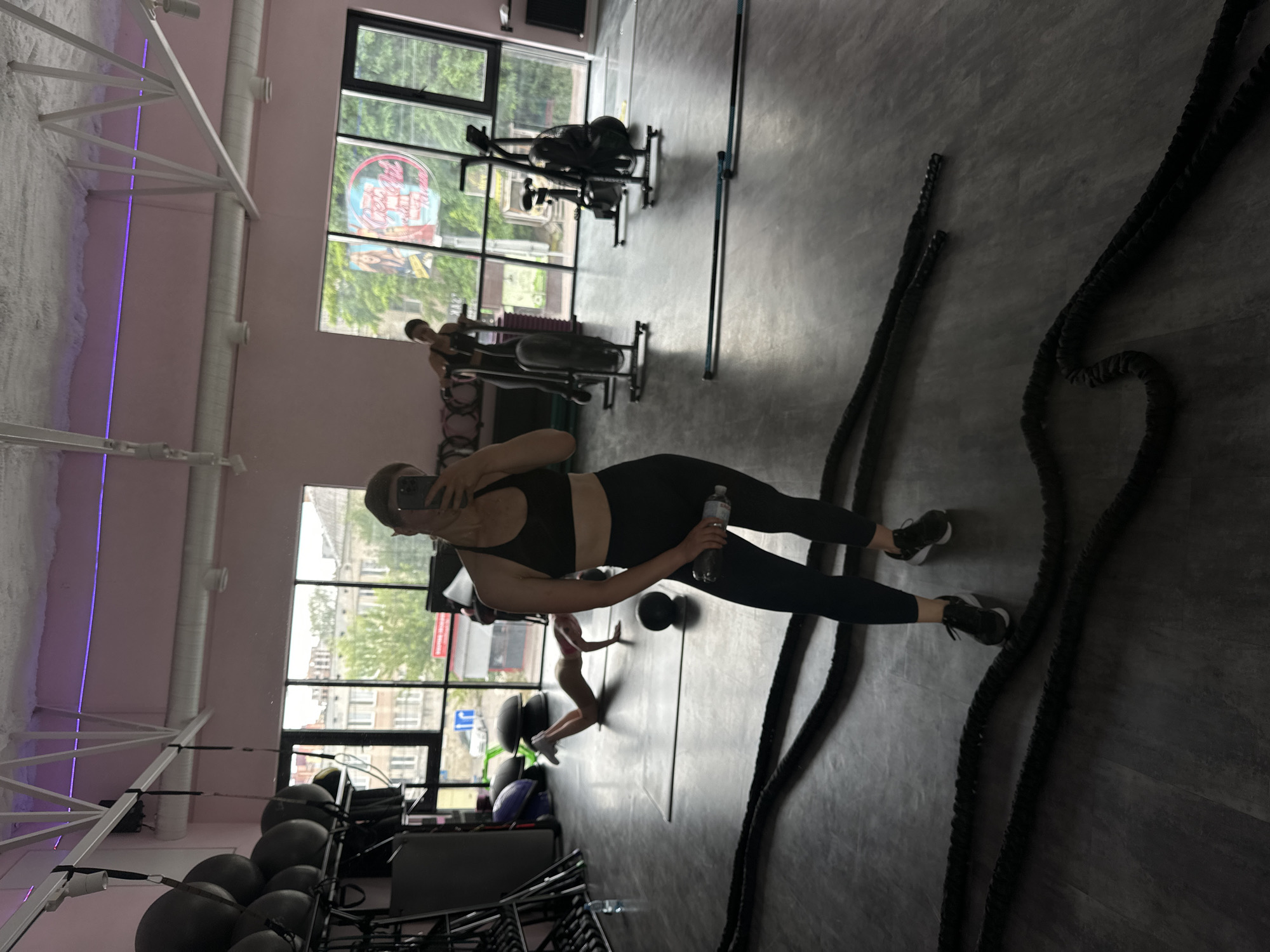 The image depicts the interior of a gym in Ternopil, Ukraine. In the foreground, a woman is taking a selfie with her phone. She is wearing a black sports bra and black leggings, and she is holding a water bottle in her right hand. Behind her, another woman is exercising on an air bike. In the background, various gym equipment is visible, including battle ropes, medicine balls, and stability balls. Large windows offer a view of the city's buildings and trees. The gym has pink walls and a white ceiling with exposed beams.