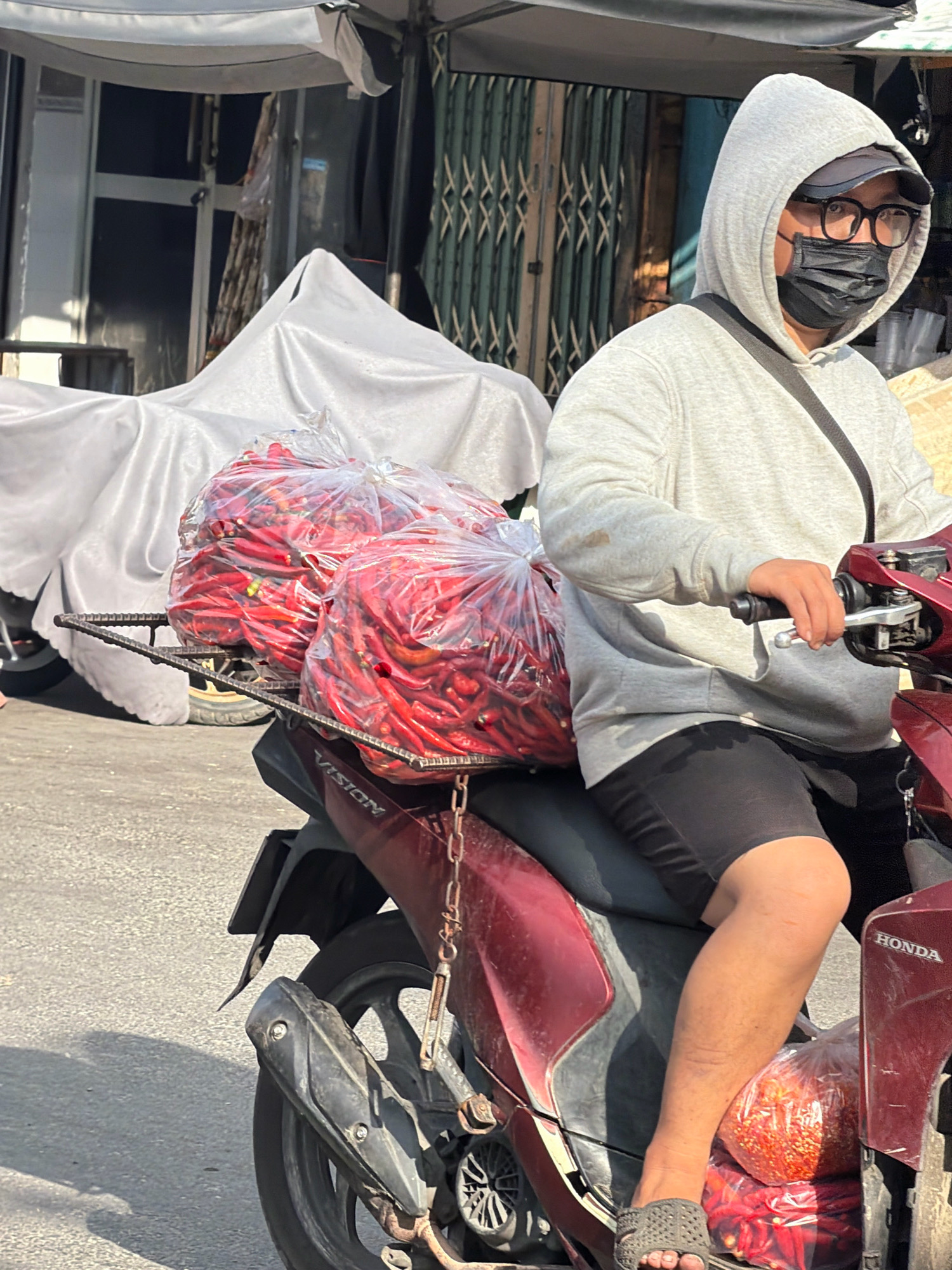 In Nha Trang, Vietnam, a person wearing a grey hoodie, glasses, and a black face mask rides a dark red Honda scooter. The scooter carries two large plastic bags filled with red chili peppers strapped to the rear. The person is wearing black shorts and a black sandal on their left foot. The background shows a street scene with market stalls and a large white tarp covering an object. The lighting suggests it is daytime, possibly morning or afternoon, with shadows cast on the asphalt road.