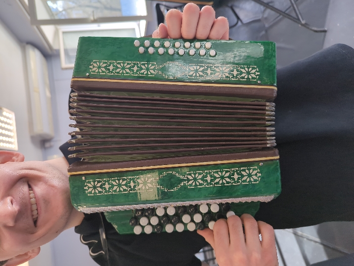 A man with a fair complexion and a visible smile holds a green bayan (button accordion) in an indoor setting. He is wearing a dark shirt. The bayan features white snowflake-like decorative patterns on its front panels, dark brown bellows, and silver-toned button mechanisms. The left side of the instrument has multiple rows of alternating black and white buttons, while the right side displays a vertical column of white buttons. The room contains an overhead rectangular fluorescent light fixture, light-colored walls, and a wall-mounted air conditioning unit. A white-framed window, partially ajar, is visible to the right, showing ambient outdoor light and blurred foliage. This scene is located within a room, possibly an institutional or educational setting, in Moscow, Russia.