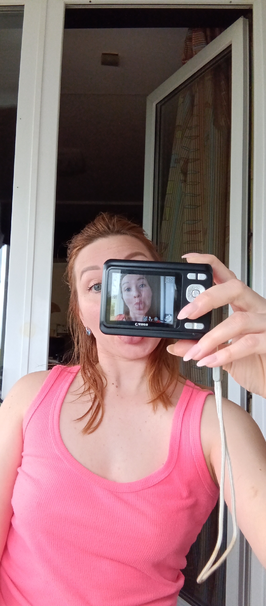 A woman with red hair and wearing a pink tank top is taking a selfie using a black digital camera. She is sticking her tongue out in the reflection on the camera screen. The background shows an open doorway or window with white frames. The location appears to be indoors, possibly a home or apartment. The city and region are not identifiable.