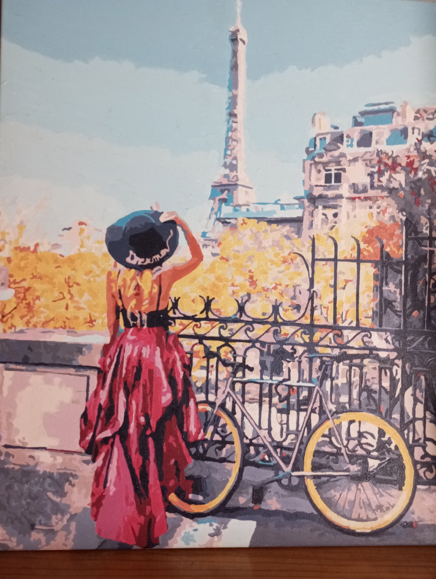 A painting depicts a woman standing on a bridge or embankment in Paris, looking towards the Eiffel Tower. She wears a wide-brimmed black hat, a black-belted top, and a full, red skirt. To her right, leaning against an ornate black iron fence, is a bicycle with yellow wheels. The background includes the Eiffel Tower, other Parisian buildings, and foliage, possibly autumn leaves. The style is impressionistic and somewhat simplified, reminiscent of paint-by-numbers. The location is Paris, France. The woman's action is looking at the Eiffel Tower.