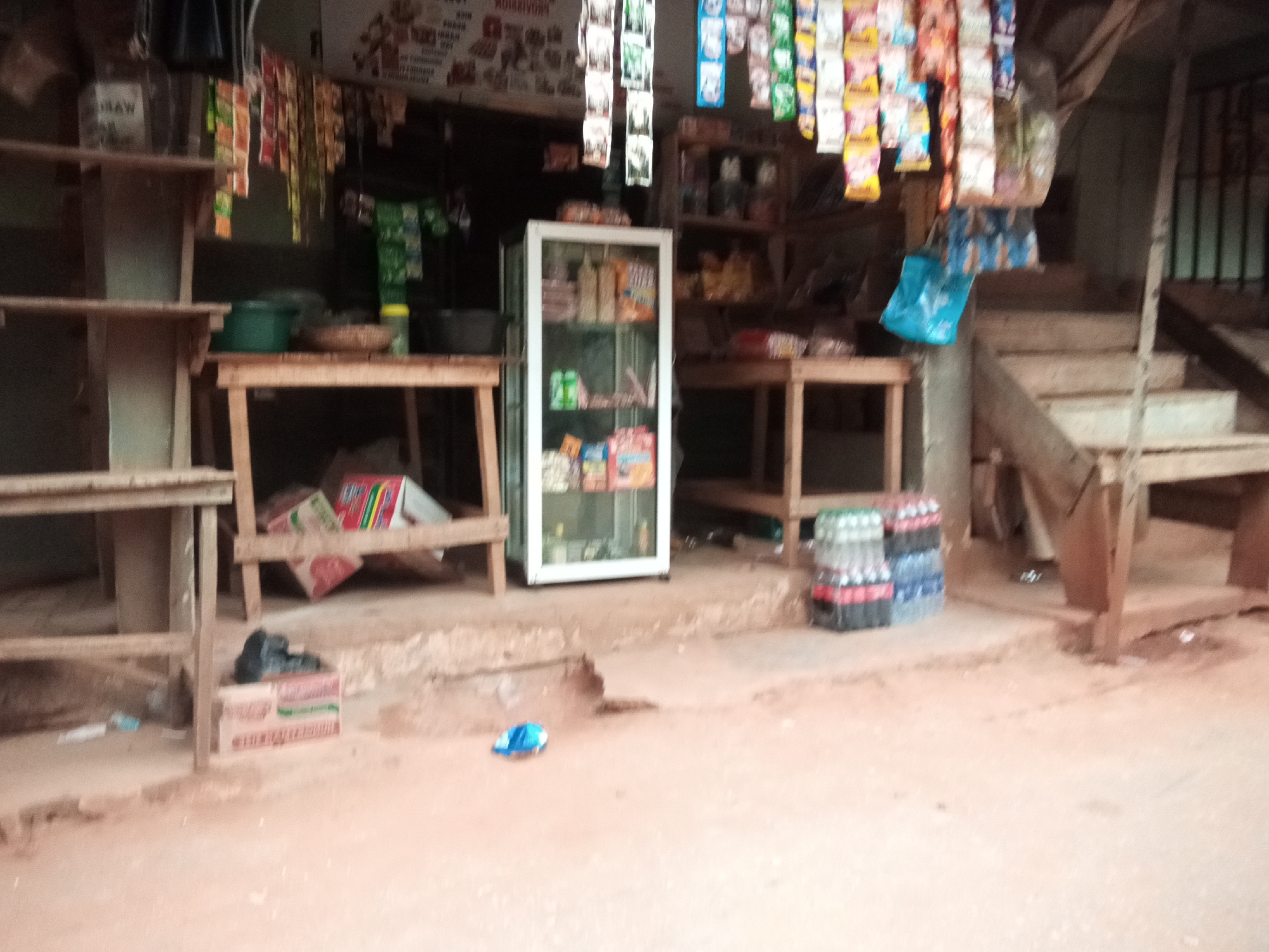 The image shows a small roadside shop in Oko Town, Nigeria, filled with various items. In the foreground, a blue packet lies on the reddish-brown ground. To the right, stacks of bottled drinks, primarily Coca-Cola and possibly Sprite, are arranged in two columns. Behind these, a wooden structure with steps leads upwards.
The shop itself features wooden shelves stocked with a variety of goods. On the left, some shelves are empty, while others hold containers, including a green bucket and a woven basket with items inside. A glass-fronted display cooler stands in the center, showcasing packaged snacks and drinks. Above the cooler, a shelf holds more packaged items. Hanging from above are numerous small, colorful packets of snacks or condiments, suspended by strings. To the far left, a white container labeled "MAW" is visible on a shelf. Further into the shop, more shelves are packed with items, and a partially visible black plastic bag rests on a cardboard box in the foreground. The overall environment appears to be dusty, with the ground covered in reddish-brown earth.