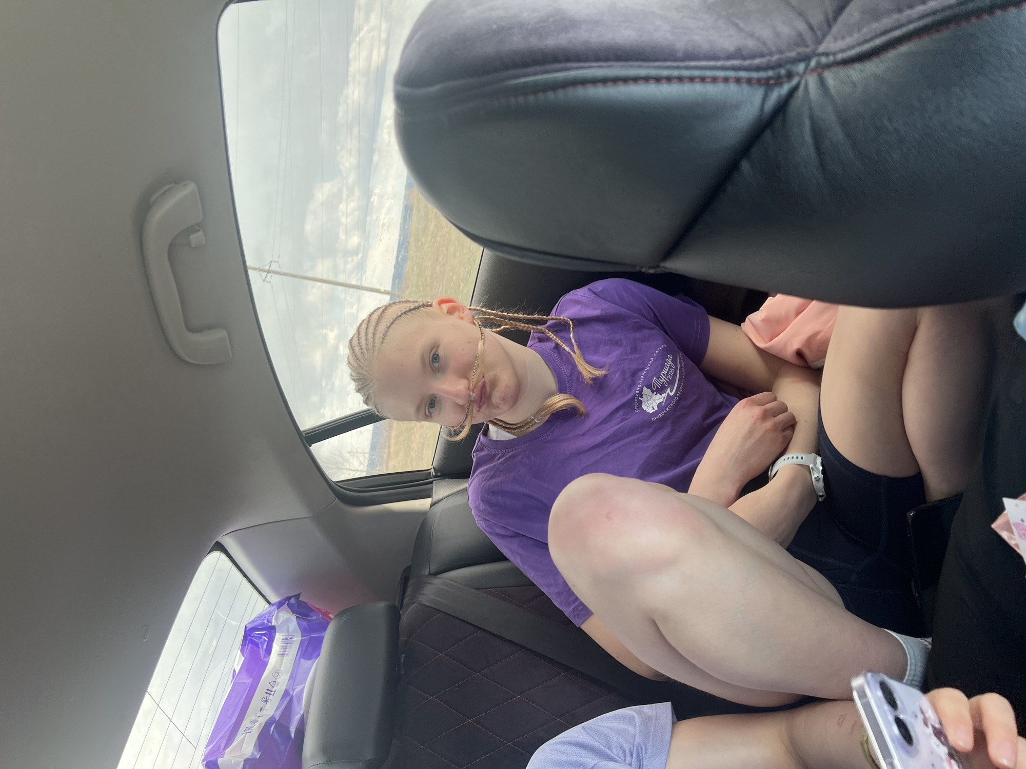 This image captures a young person seated in the back of a car, likely during daytime under an overcast sky. The primary subject, a person with blonde hair styled in cornrow braids, is wearing a purple short-sleeved t-shirt and dark shorts, looking directly at the camera with a neutral expression. A white wristband is visible on their left arm. The purple t-shirt features Cyrillic text, partially visible as "СПОРТИВНО-ТВОРЧЕСКИЙ ЛАГЕРЬ ПУРНАГА 2023", which translates to "SPORTS AND CREATIVE CAMP PURNAYA 2023", aligning with the given Malaya Purga, Russia location context.
The setting is the interior of a car, with dark leather seats (the front headrest is prominent) and a patterned fabric back seat. A white grab handle is visible on the ceiling. Through the car windows, a cloudy sky and distant power lines or poles are visible, indicating an open, possibly rural landscape outside.
In the bottom right, parts of another person's leg and arm are visible, holding a smartphone in a clear, patterned case. A large purple plastic bag with white writing, possibly packaging for goods, rests on the back seat or floor by the rear window. The activity appears to be casual travel or a break during a journey. The scene suggests a relaxed moment inside a vehicle, possibly on the way to or from the sports and creative camp mentioned on the t-shirt.