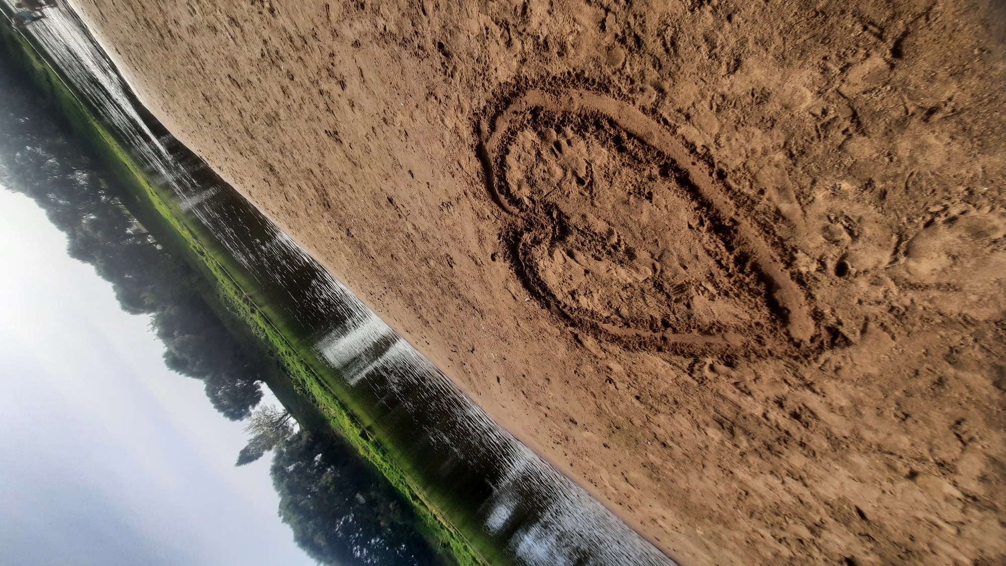 The image captures a scenic view of a riverbank, possibly in Shklov, Belarus. In the foreground, a heart shape is drawn in the wet sand, indicating human presence and activity. The river, with its dark, reflective water, flows gently, bordered by lush green vegetation and a line of trees. The sky is bright, suggesting a sunny day. The overall impression is one of tranquility and natural beauty, with a touch of personal expression from the heart drawn in the sand.