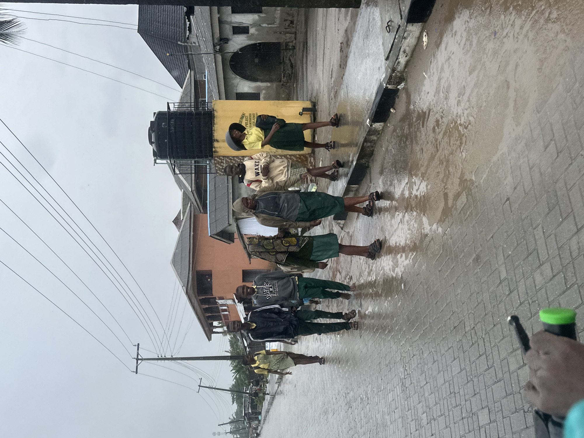 In a street scene in Badagry, Nigeria, on a rainy day, a group of young people are walking along a wet, paved road. The sky is overcast, and the road is covered in puddles, indicating recent or ongoing rainfall. In the background, buildings of various styles are visible, including residential structures and what appears to be a commercial building with a yellow facade. A large black water tank is mounted on top of one of the buildings. Utility poles and overhead wires are also present, typical of urban infrastructure. The individuals in the foreground are dressed in what seem to be school uniforms, with some wearing jackets and skirts, and others in trousers. One young woman is wearing a wide-brimmed hat. The overall impression is of a community navigating daily life under challenging weather conditions.
