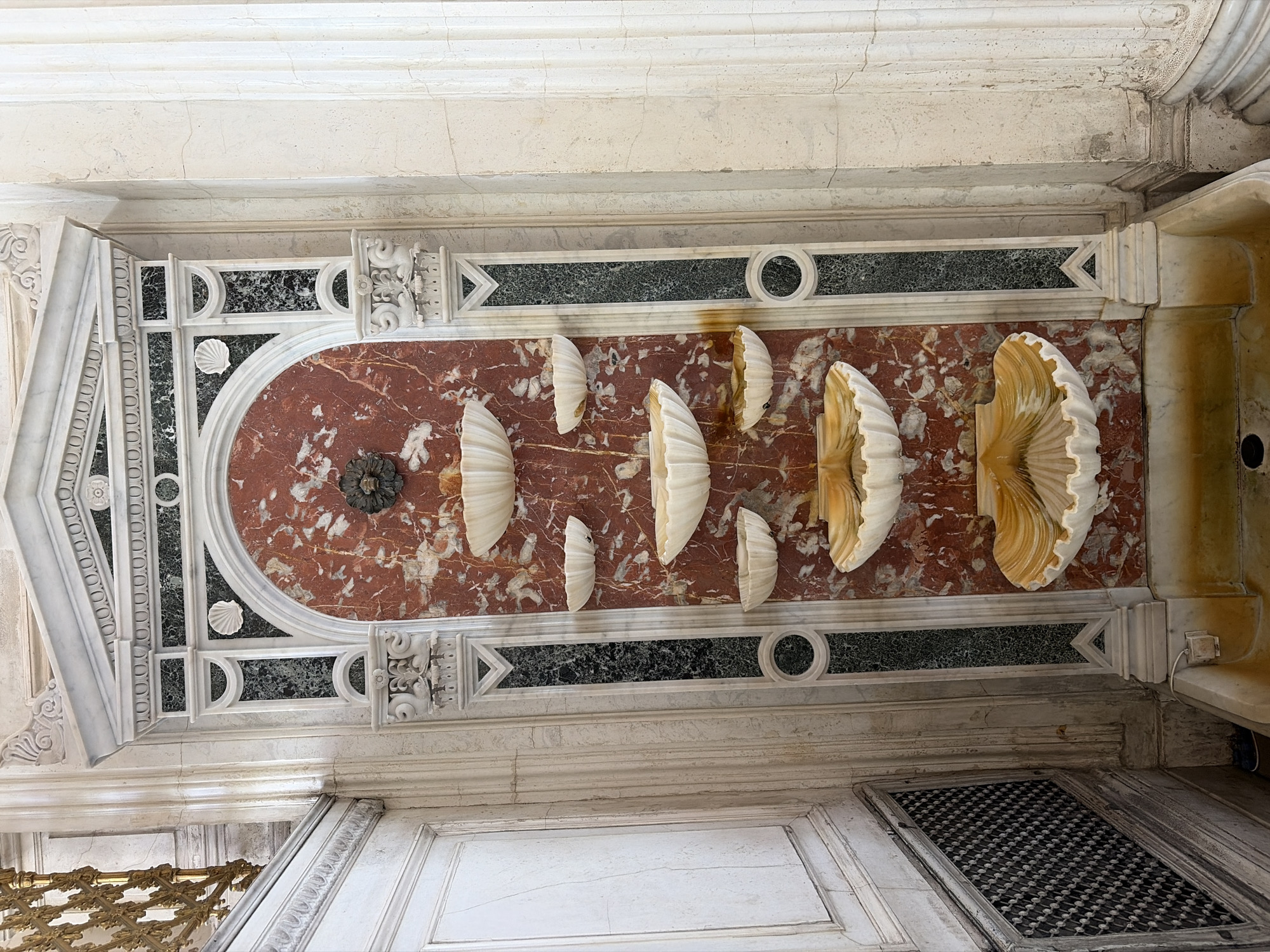 This image depicts a baptismal font in the Church of St. Catherine in Saint Petersburg, Russia. The font is decorated with multiple seashell-shaped basins arranged in a row on a marble surface with red and white veins. The surface is surrounded by white marble architecture with green and black accents. The image is taken from below.