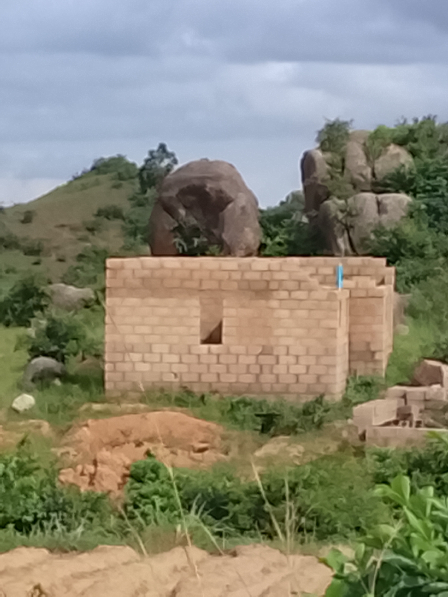 In Rafin Bauna, Nigeria, an unfinished brick building stands amidst a rugged, natural landscape. The structure, constructed from tan-colored bricks, has visible openings for windows and appears to be in an early stage of construction, with no roof or interior finishing. Surrounding the building is dense green vegetation and scattered large rocks, some of which are quite prominent and seem to be part of the natural terrain. In the background, rolling hills covered in grass and dotted with trees rise towards a cloudy sky. The scene suggests a remote or developing area where construction is taking place in a natural environment.