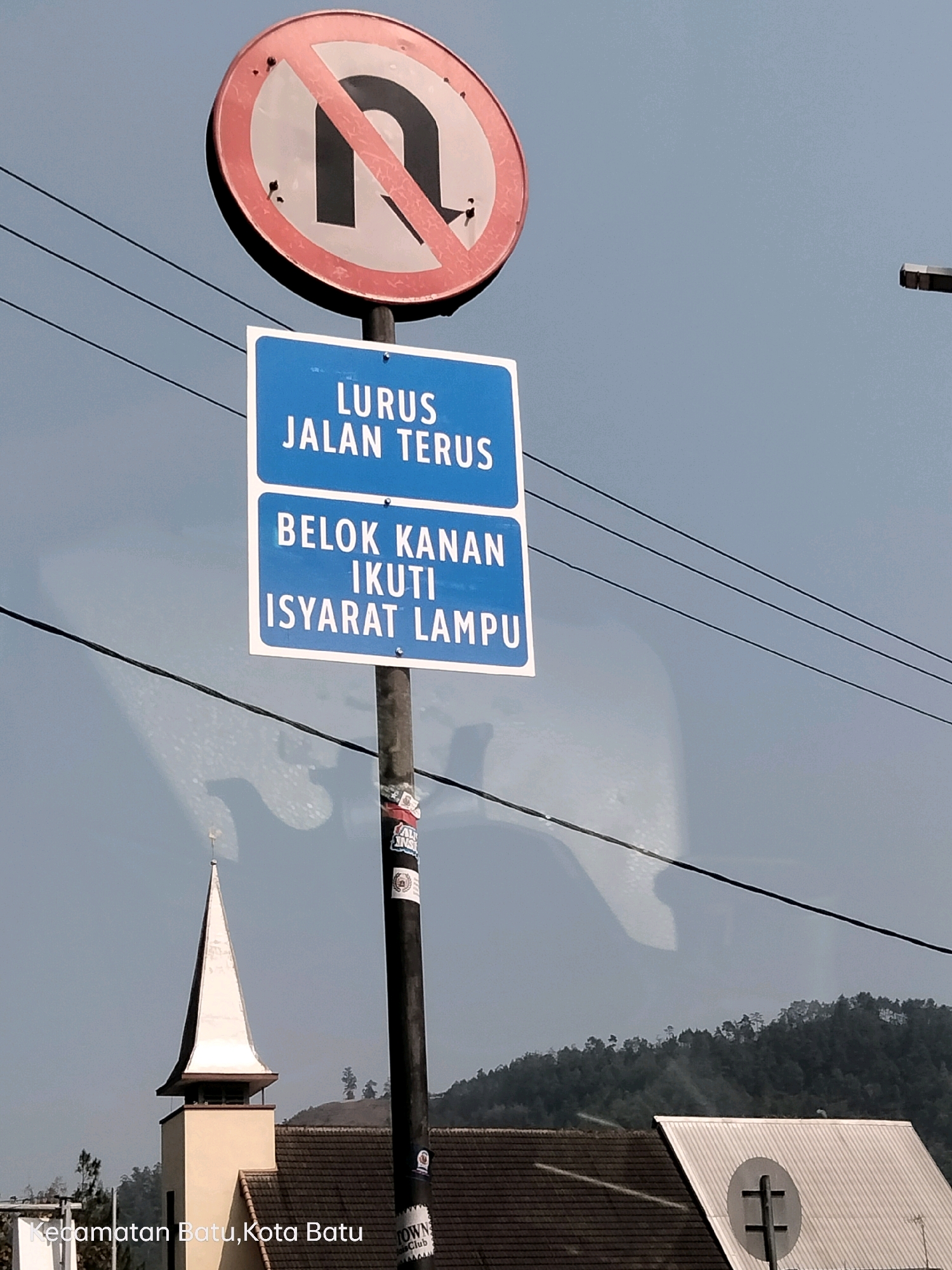 The image shows a traffic signpost with multiple signs against a clear sky. The topmost sign is a red circle with a diagonal line through a black U-turn symbol, indicating "No U-turn." Below this, two blue rectangular signs are stacked. The upper blue sign reads "LURUS JALAN TERUS" in white text, which translates to "Go Straight." The lower blue sign reads "BELOK KANAN IKUTI ISYARAT LAMPU," meaning "Turn Right Follow Traffic Signals." The signpost is a dark pole with stickers attached to it. In the background, a white-steepled building, possibly a church, is visible with a tiled roof. Beyond that, a forested hill rises. The text overlay at the bottom of the image identifies the location as "Kecamatan Batu, Kota Batu," which is Batu District, Batu City, Indonesia. Electrical wires run across the upper portion of the frame.