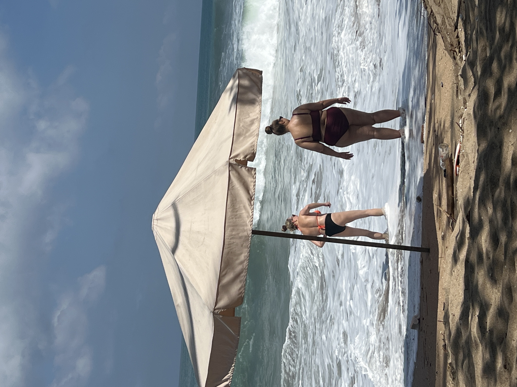 Two women stand in shallow ocean water near the shore under a large, tan beach umbrella. The woman on the left wears a black bikini with an orange top. The woman on the right wears a maroon two-piece swimsuit. Both women are facing the ocean, with large waves breaking behind them. The water is frothy and white. The beach in the foreground is sandy with some scattered debris. The sky is blue with some clouds. This scene is consistent with the beaches in Kuta, Indonesia, known for its surfing and beach activities.