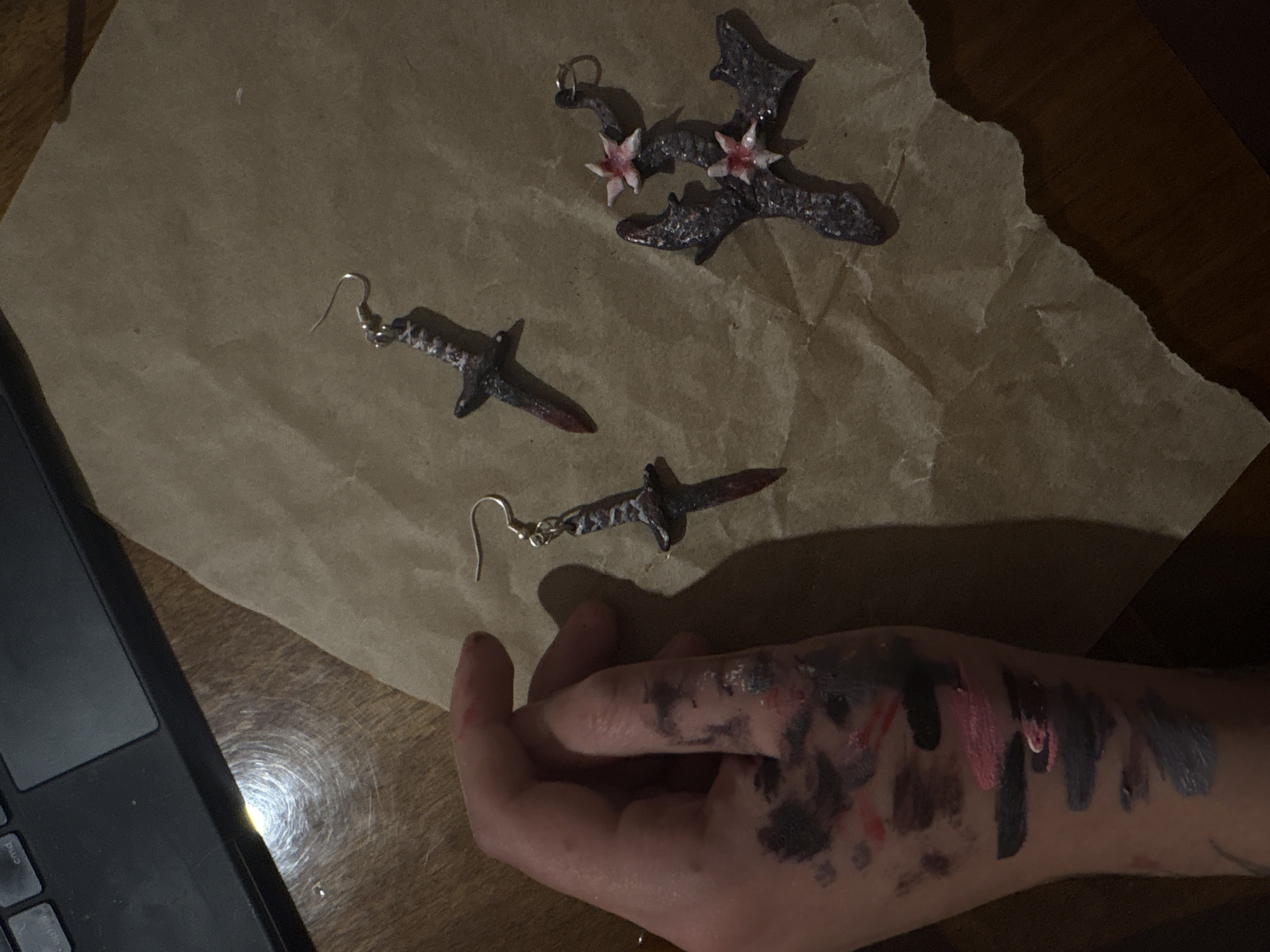 A low-angle shot depicts a collection of dark-colored, crafted jewelry pieces arranged on a crumpled piece of brown paper. To the left is part of a black laptop. At the lower right is a hand and lower arm with streaks of dark paint across the skin. There is no indication of the city Yerevan, Armenia.