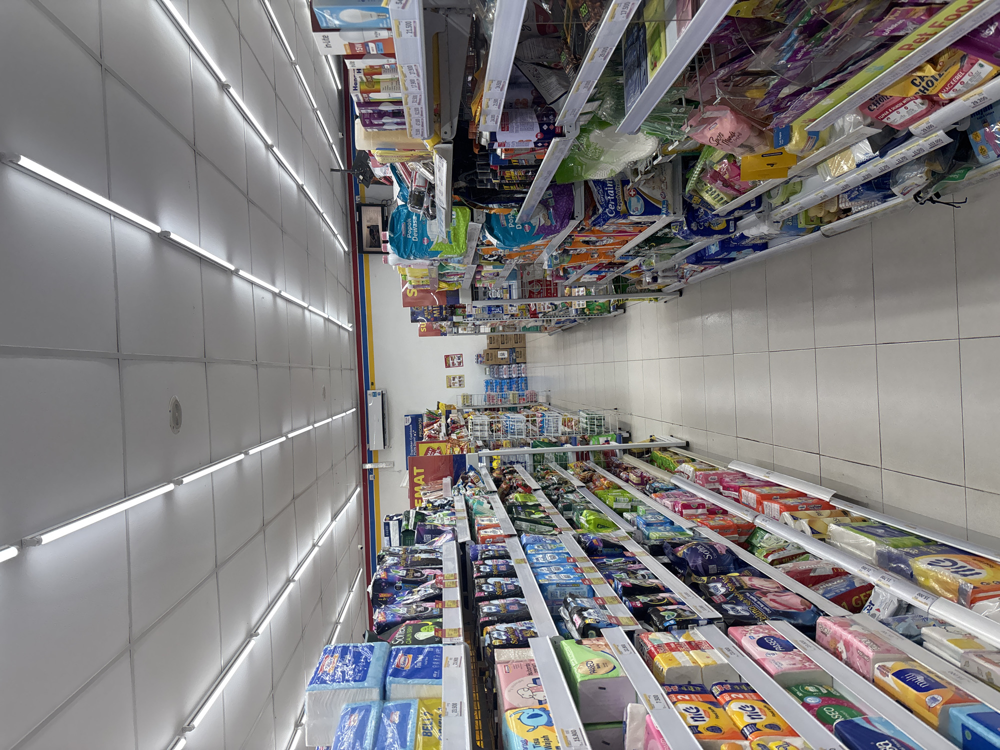 The image depicts the interior of a brightly lit convenience store, likely a minimarket, in Cirebon, Indonesia. The perspective is from within an aisle, looking down a long corridor lined with shelves stocked with various consumer goods. The ceiling features a grid of white tiles with rows of fluorescent tube lights illuminating the space. The floor is tiled with large, light-colored square tiles.
On the left side of the aisle, shelves are filled with packaged items such as facial tissues, sanitary napkins, diapers, and other personal care products. Many packages display prices, with denominations like "23.500" and "15.900" visible. Brands like "Paseo," "Nic," and "Multi" are identifiable. Further down this aisle, more rows of merchandise are visible.
The right side of the aisle also displays shelves packed with products. This side appears to feature a wider variety of items, including cleaning supplies, pet food, and potentially beverages or snacks. Some labels include "WHISKAS," "TUNA & SALMON," and "MACKEREL." Prices are also visible on this side, such as "29.000" and "17.500."
In the background, the store continues with more aisles and displays. A sign with "PROMOSI" suggests promotional offers are available. There is also a wall-mounted air conditioning unit visible on the right side. The overall impression is a well-organized retail environment typical of a convenience store in urban Indonesia. No people are present in the immediate scene, and no specific actions or interactions are captured. The lighting is even and bright, indicating it is likely daytime or the store is well-illuminated for operations.