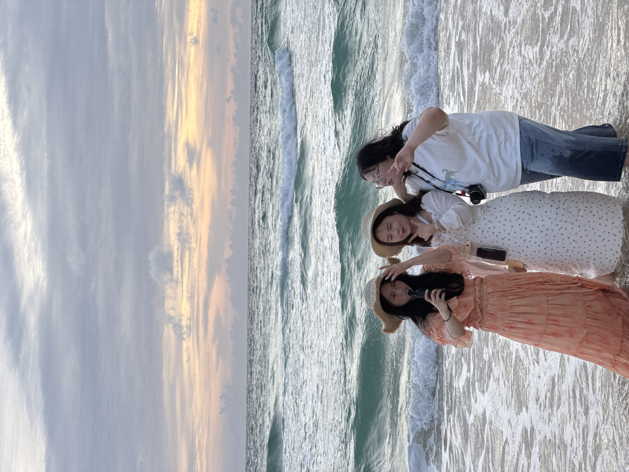 Three young women are standing in the shallow surf on Karon Beach, Thailand. The sky is cloudy with streaks of pink and yellow from the setting sun. The ocean is a vibrant turquoise with white-capped waves rolling in.
The woman on the left is wearing a flowy peach-colored dress and a straw hat. She is holding a black camera or phone and appears to be taking a selfie. The woman in the middle is wearing a white dress with a blue floral pattern and a straw hat. She is making a peace sign with her fingers. The woman on the right is wearing a white t-shirt and denim jeans. She has glasses and is also making a peace sign with her fingers. She has a camera hanging around her neck. The women are all looking towards the camera.
