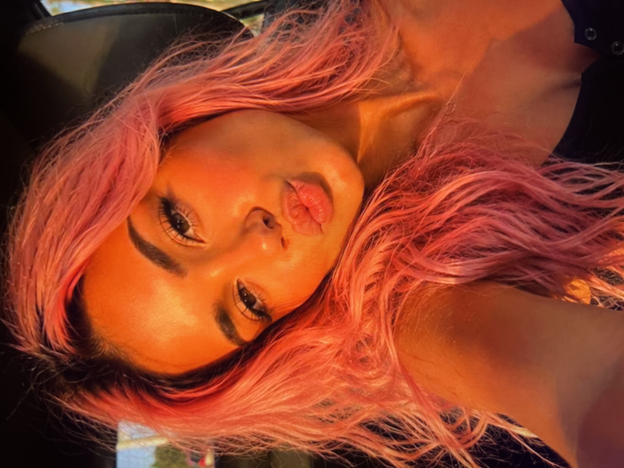 The image is a close-up selfie of a young woman with bright pink hair, taken from the chest up, with her head tilted and mouth pursed in a kiss. The lighting suggests it was taken outdoors during golden hour, casting a warm, orange glow over her face and hair. She appears to be in a car, with the dark interior visible in the background and some blurred greenery or sky suggesting an external environment. Her makeup is visible, with defined eyebrows and mascara.