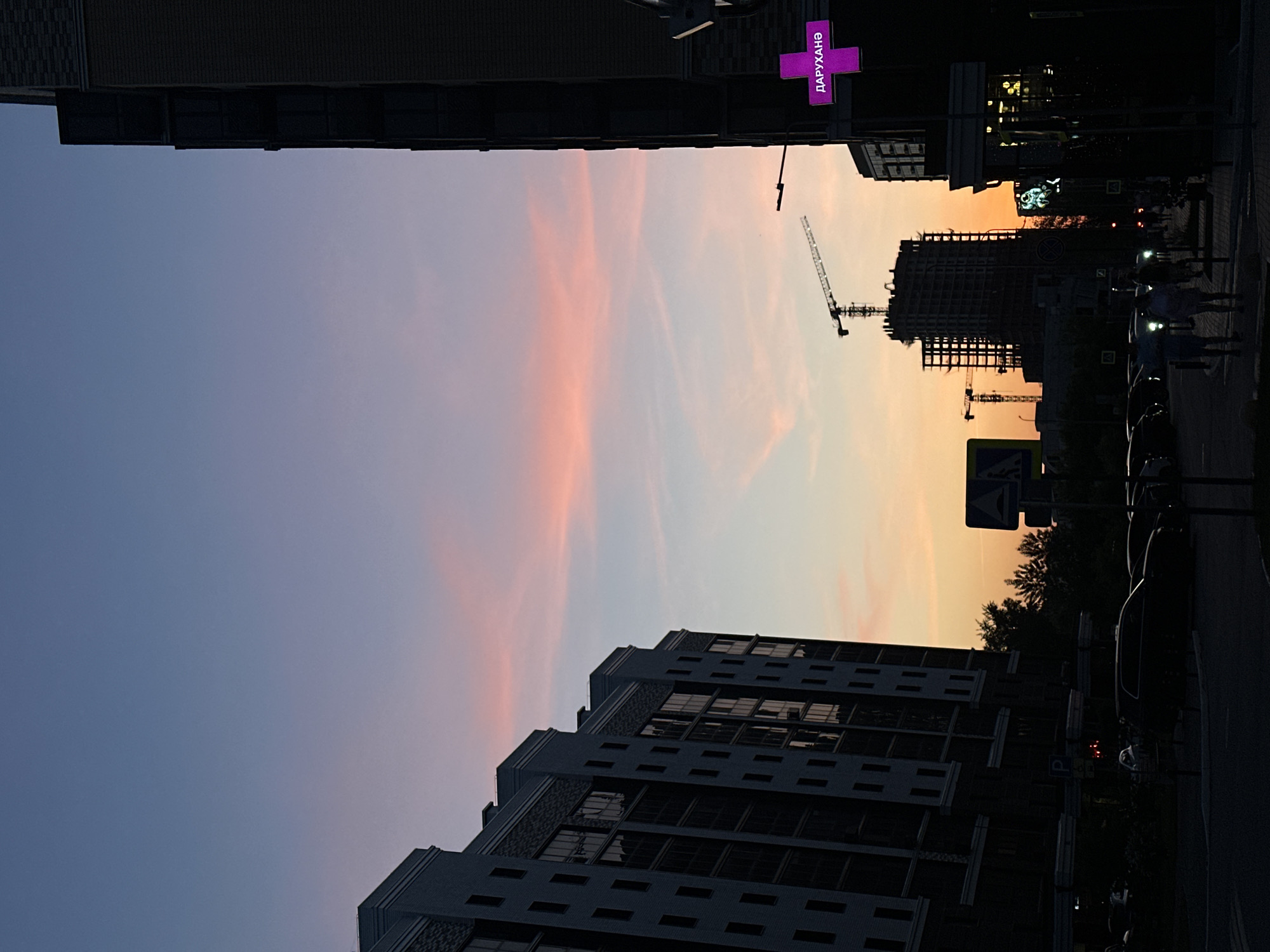 The image captures a street scene in Kazan, Russia at dusk. The sky transitions from blue to orange and pink. Silhouetted buildings frame the sky, with a modern apartment building on one side and a construction site with cranes on the other. A purple pharmacy sign with Cyrillic text is visible near the construction site. Pedestrians and parked cars line the street, and street signs are posted.