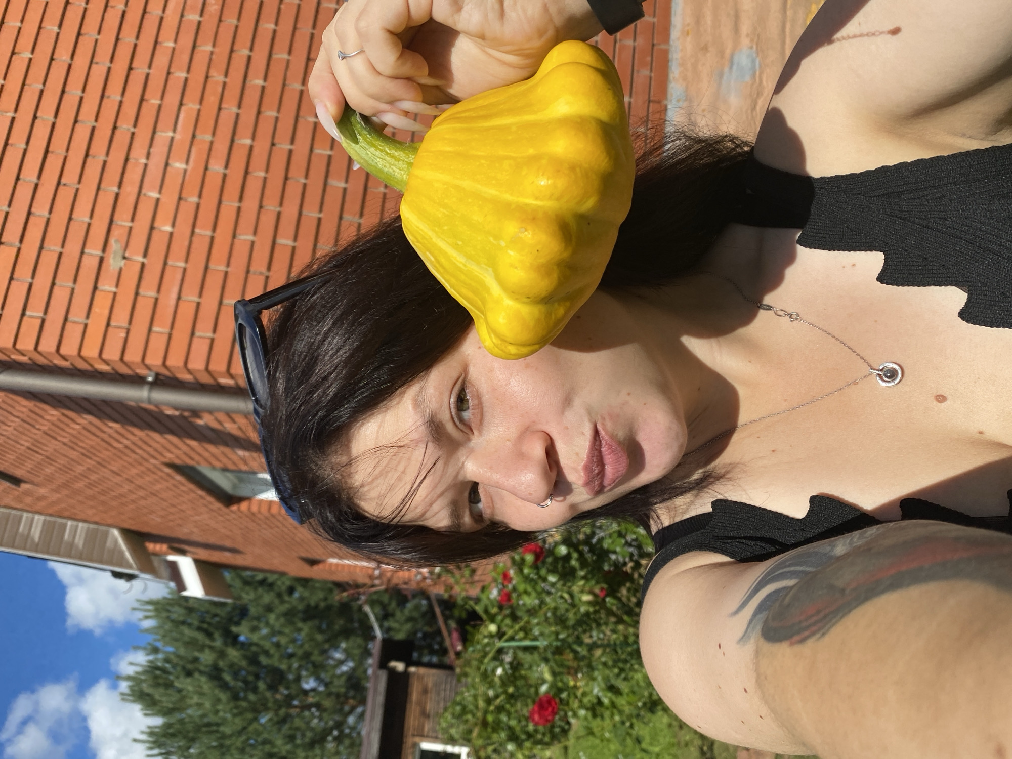 The image shows a young woman holding a yellow pattypan squash in front of a brick building and greenery. The woman has dark hair and is wearing a black top. She is outdoors on a sunny day. The location is identified as Fokino, Russia. The scene suggests a rural or suburban setting, possibly a backyard or garden, given the presence of the squash and the vegetation. The brick building is a prominent feature in the background.