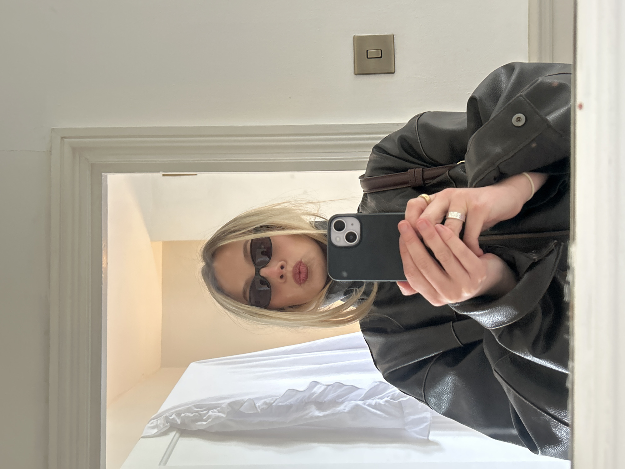 A young woman is taking a mirror selfie. She wears sunglasses and a dark, oversized leather jacket. She holds a smartphone with a dark case in her hands. Her facial expression is a slight pout. She has fair skin and blonde hair. She wears rings on her fingers. The background is a white or light-colored room with a partially visible bed with white sheets. A light switch is on the wall above her right shoulder. The scene is likely an interior setting in a residential dwelling. Location information is not discernible from the image.