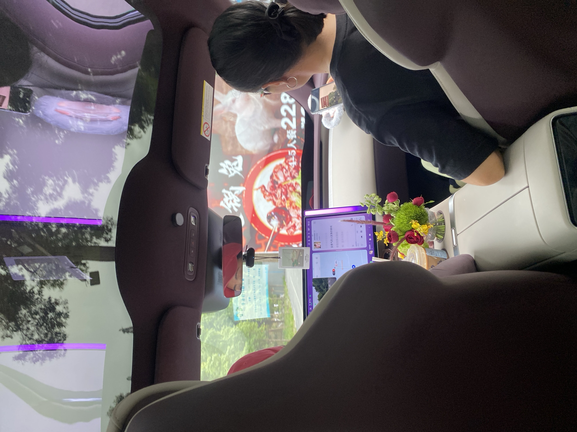This image, taken in Chongzhou City, China, shows the interior of a vehicle, likely a car, from the perspective of a passenger. The scene is dominated by the car's interior features, including the seats and dashboard. In the foreground, a person is seated, looking towards the right, with their head and shoulders visible. They appear to be wearing a dark shirt and have their hair pulled back.
On the dashboard, a screen displays an advertisement for a meal set for "5 people, 228" (presumably in Chinese Yuan). The text "没勇气的人就像的瞬间" is also visible on the screen, which translates to "People without courage are like moments." Adjacent to this screen is a tablet or secondary display showing what appears to be a map or navigation interface with some Chinese text, including icons for music playback. A small vase with a bouquet of red and yellow flowers and a green floral arrangement sits on a surface near the screen.
The car's interior is upholstered in a dark color, possibly maroon or dark purple, with white trim. The windshield reflects the outside environment, showing trees and a bright sky, suggesting it's daytime. The overall mood is casual, perhaps a passenger in a ride-sharing service or a private car on a journey. The presence of a screen displaying advertisements and entertainment, along with a navigation system, points to a modern vehicle interior.
