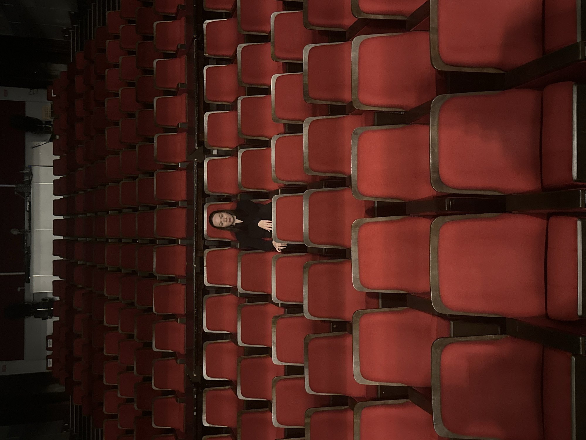An overhead photograph captures a vast, dimly lit auditorium primarily filled with numerous rows of empty, red theater seats. Each seat features a plush red cushion, dark armrests, and metallic accents on its frame. The seats are arranged in a structured grid, receding into the distance, creating a sense of depth and scale.
A single individual is positioned in one of the central rows, lying horizontally across two adjacent seats. The person has dark hair and is wearing a black long-sleeved garment. They are looking directly upwards towards the camera with a neutral, calm expression. Their arms are resting across their body.
The lighting in the auditorium is muted, allowing the vibrant red of the seats to stand out against the darker surrounding elements, such as the shadowy area to the left which likely represents the stage or front of the hall. The scene suggests a quiet, unoccupied venue, highlighting the contrast between the multitude of empty seats and the solitary presence of the person. No text is visible in the image.