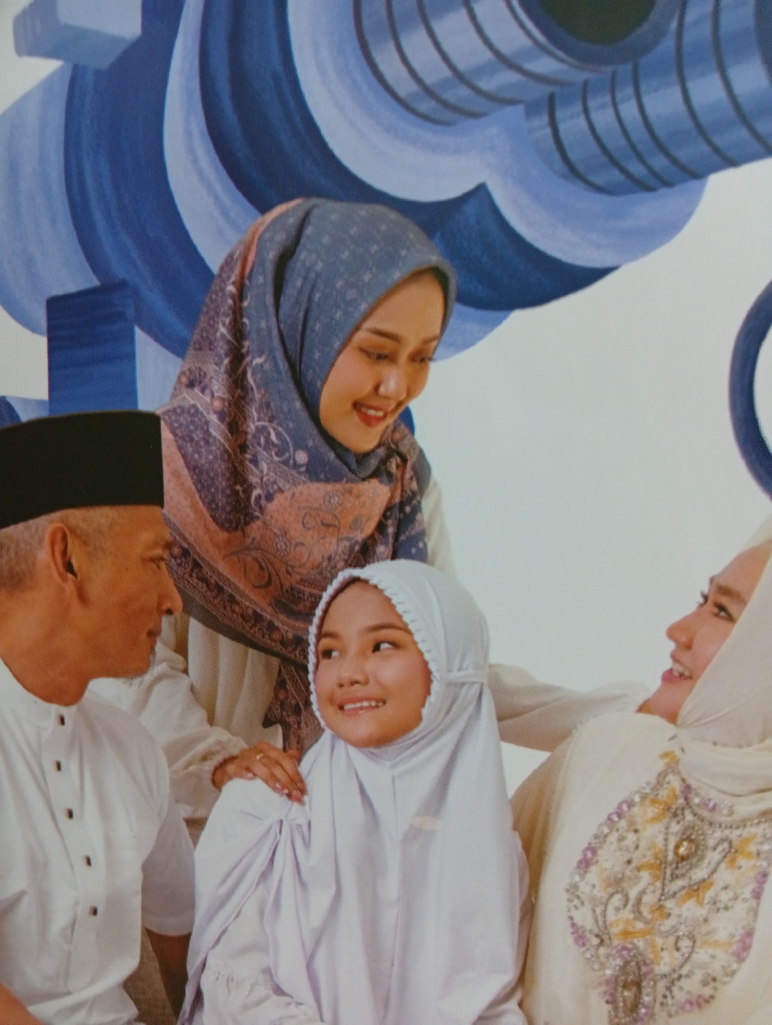 The image shows a portrait of a family in Tlogomulyo, Indonesia. The background features a mural with abstract blue and white wavy patterns. In the foreground, four individuals are visible. On the left, an elderly man wears a black peci hat and a white collared shirt with dark buttons. Next to him, a younger woman with a blue and pink patterned hijab smiles and looks down. In the center foreground, a young girl wears a white hijab and white clothing, looking to her left. To the right, another woman, partially visible, wears a light-colored hijab and a cream-colored garment with ornate embroidery. The people are arranged closely, suggesting a family gathering or photoshoot.