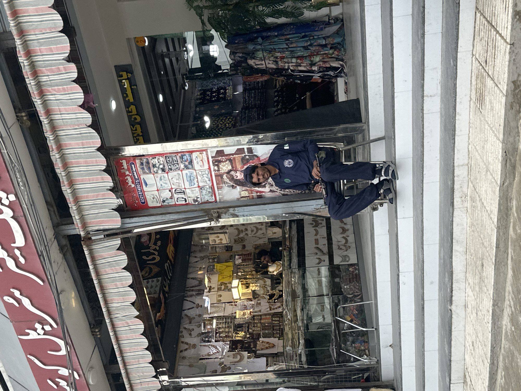 The image shows a street scene in an urban environment. A man in a police uniform is standing in front of a shop. The shop has signs in Arabic script and displays various items inside. The scene is partially obscured by awnings and structural elements. The background features other shops and a building with vertical columns.