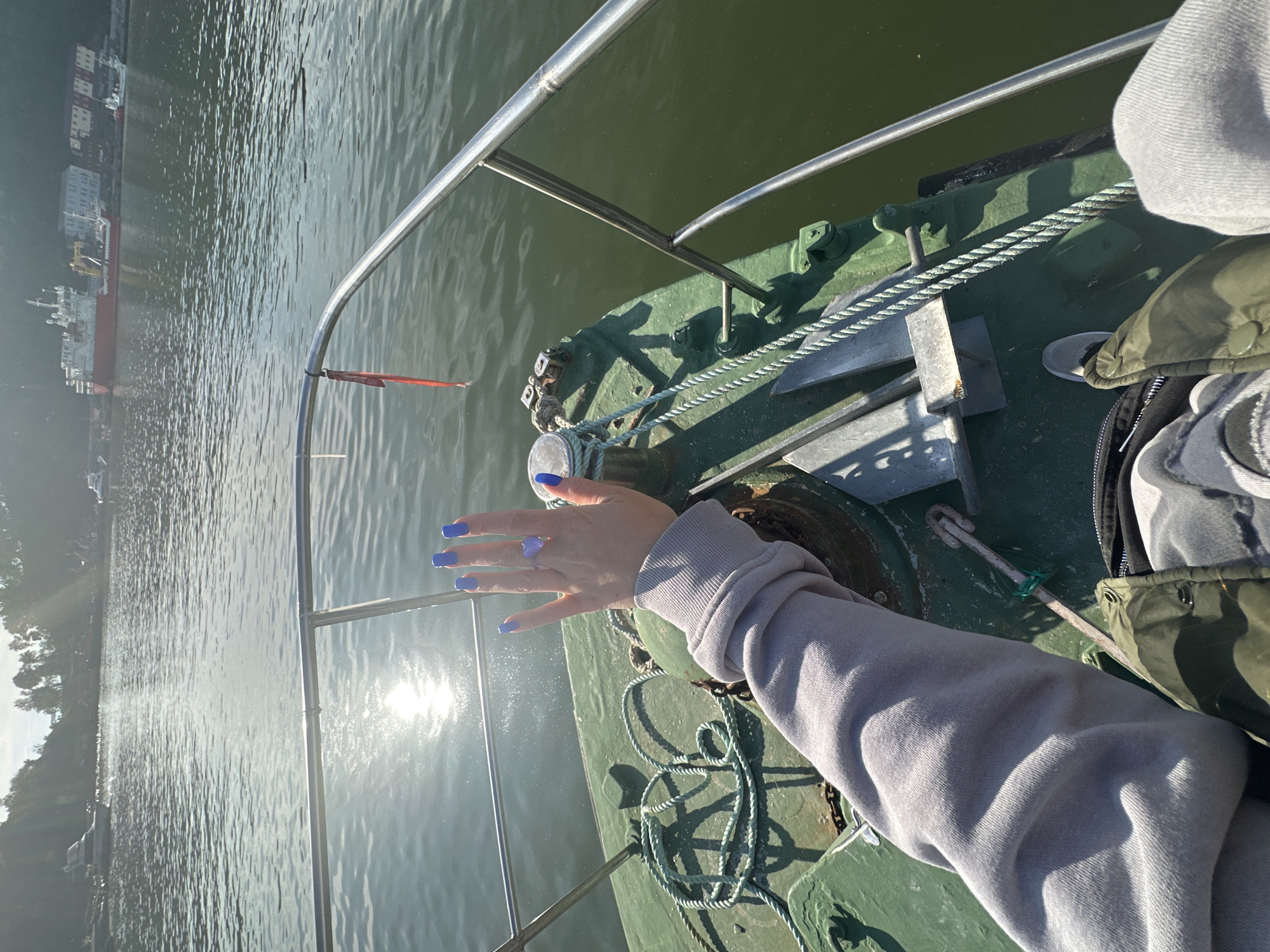 A first-person perspective shot is taken from the deck of a green boat navigating through water, with the sun casting a strong glare on the surface. The boat's metallic railing is visible in the foreground. In the middle ground, a person's hand with bright blue fingernails and a heart-shaped ring is extended, resting on the boat's deck. The deck itself is green and features a coiled rope and a metal cleat. Further out in the water, several boats and a red cargo ship are visible near the shore, with buildings lining the coast. Trees are also present on the elevated terrain. The overall scene suggests a sunny day spent on the water near a coastal city. The location is implied to be Petropavlovsk-Kamchatsky, Russia, given the context.