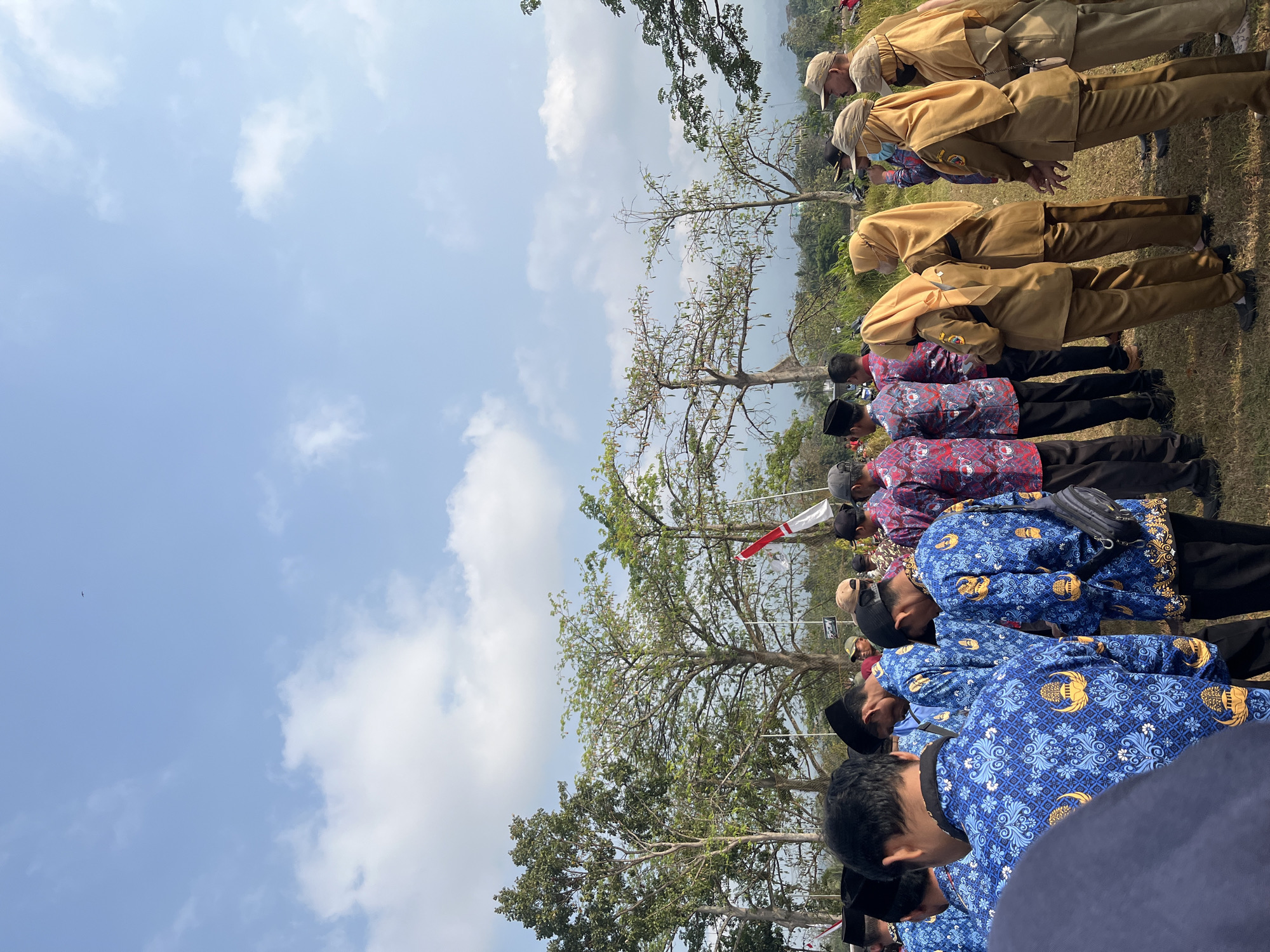 The image depicts a gathering of people in Gembong, Indonesia, on a sunny day. The sky is a clear blue with scattered white clouds, and a single bird is visible in the distance. The foreground is dominated by a group of individuals, most of whom are wearing blue batik shirts with intricate patterns of yellow and white motifs, and some are wearing black hats or caps. Behind them, a line of people in tan uniforms stands, some wearing headscarves. Trees with green foliage and bare branches are present in the middle ground, and a red and white Indonesian flag is visible. The overall scene suggests an outdoor event or ceremony.