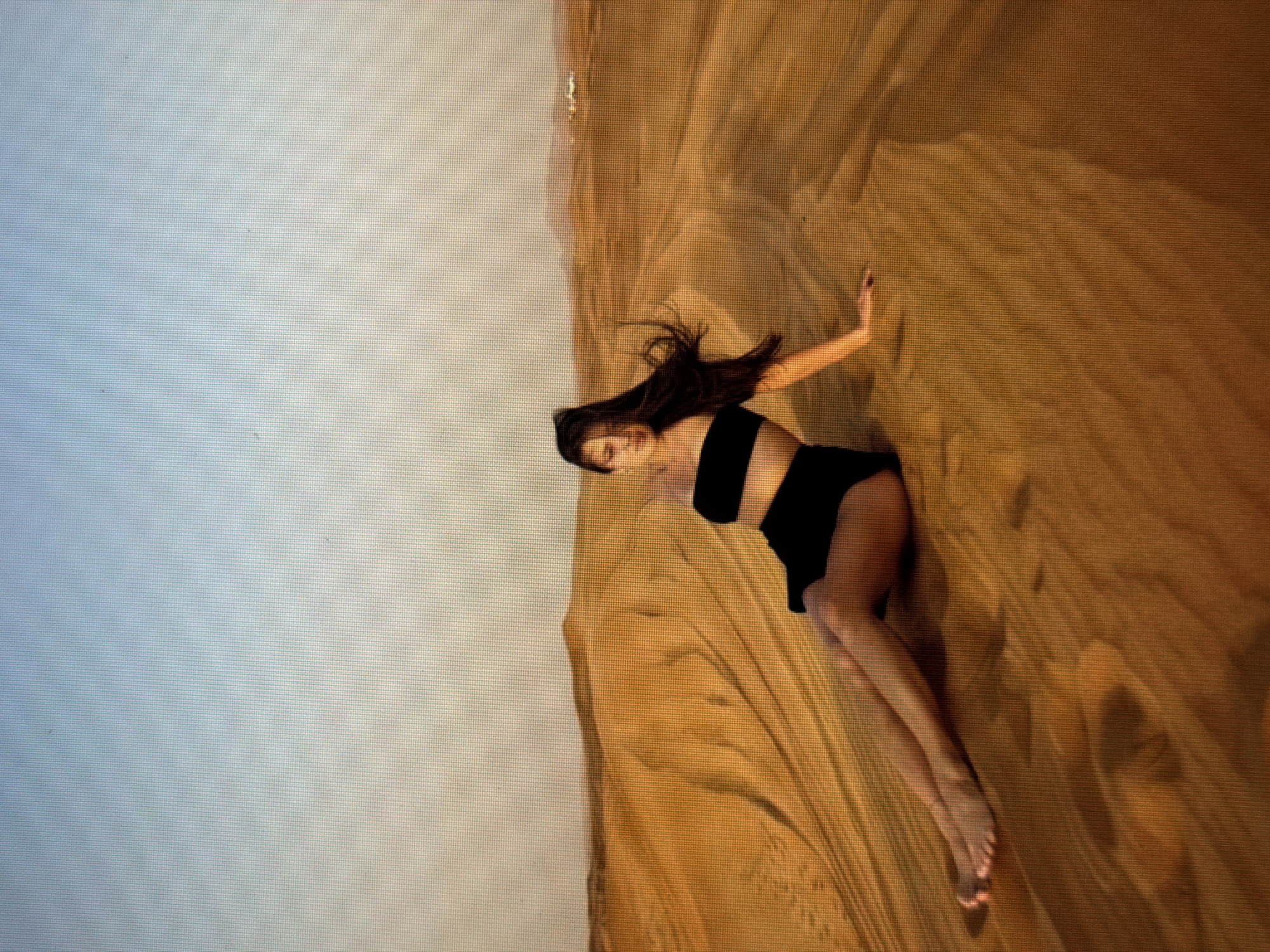 A young woman with long, dark hair lies on sand dunes, her body tilted at an angle as if caught in a moment of playful movement. She is wearing a black two-piece outfit. Her left arm is extended upwards, reaching towards the rippling sand, while her right arm is bent, her hand resting near her head. Her legs are also extended, one bent at the knee, showcasing her bare feet. The sand dunes are a warm, golden brown, with visible textures and shadows indicating the contours of the landscape. The sky above is a gradient of pale blue and white, suggesting a clear day. The scene appears to be a desert environment. The low angle of the sun, casting long shadows, hints at either early morning or late afternoon. The overall mood seems relaxed and serene. No visible text is present in the image.