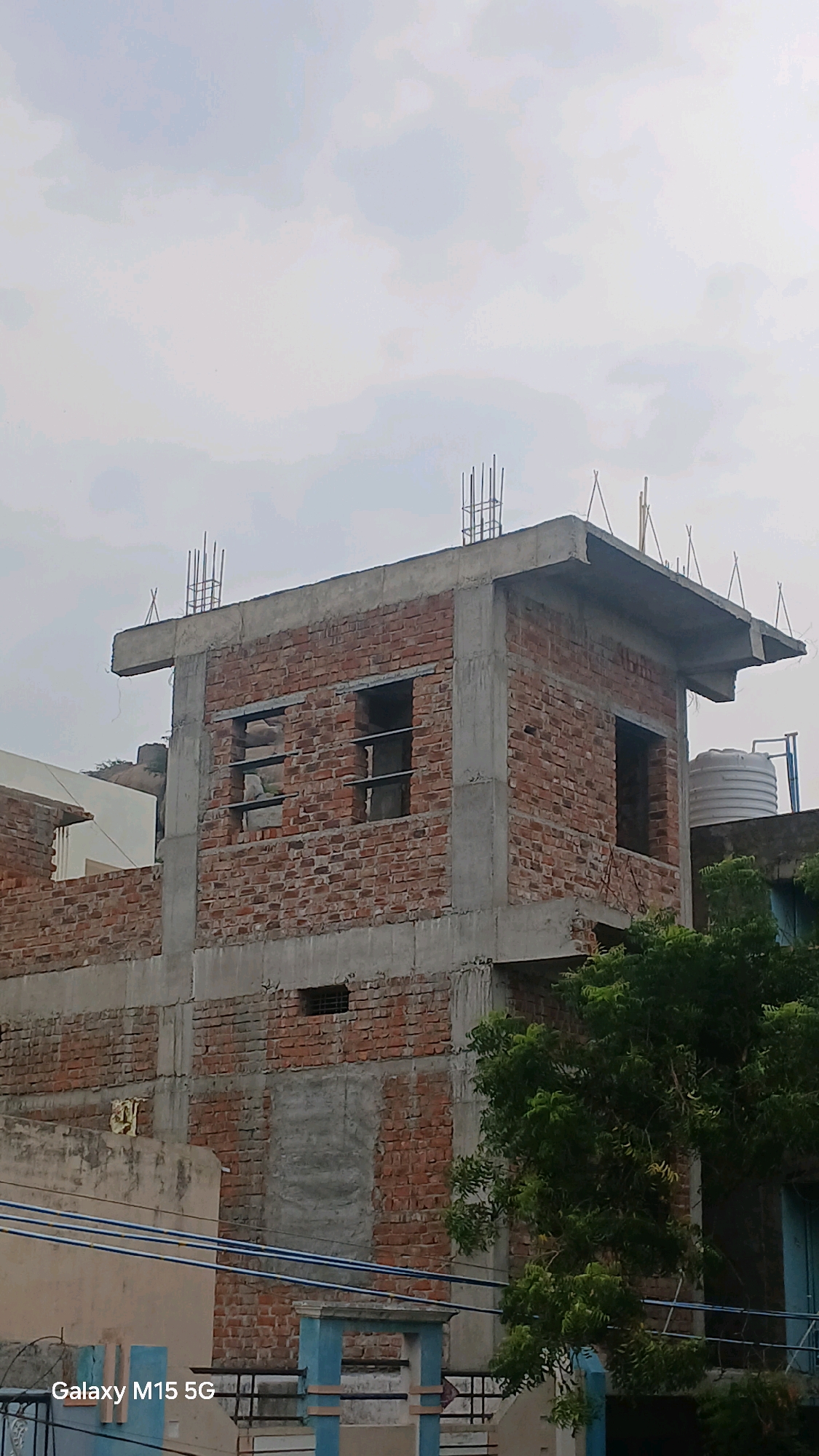 The image shows an unfinished brick building under construction in Uravakonda, India. The building has exposed concrete pillars and lintels, with brick walls partially constructed. There are openings for windows and doors. Rebar protrudes from the top of the building, indicating it is still in the early stages of construction. A large white water tank is visible on a nearby roof, and green trees obscure parts of the building. The sky is overcast with clouds. The text "Galaxy M15 5G" is visible in the bottom left corner of the image, indicating the device used to capture the photo.
