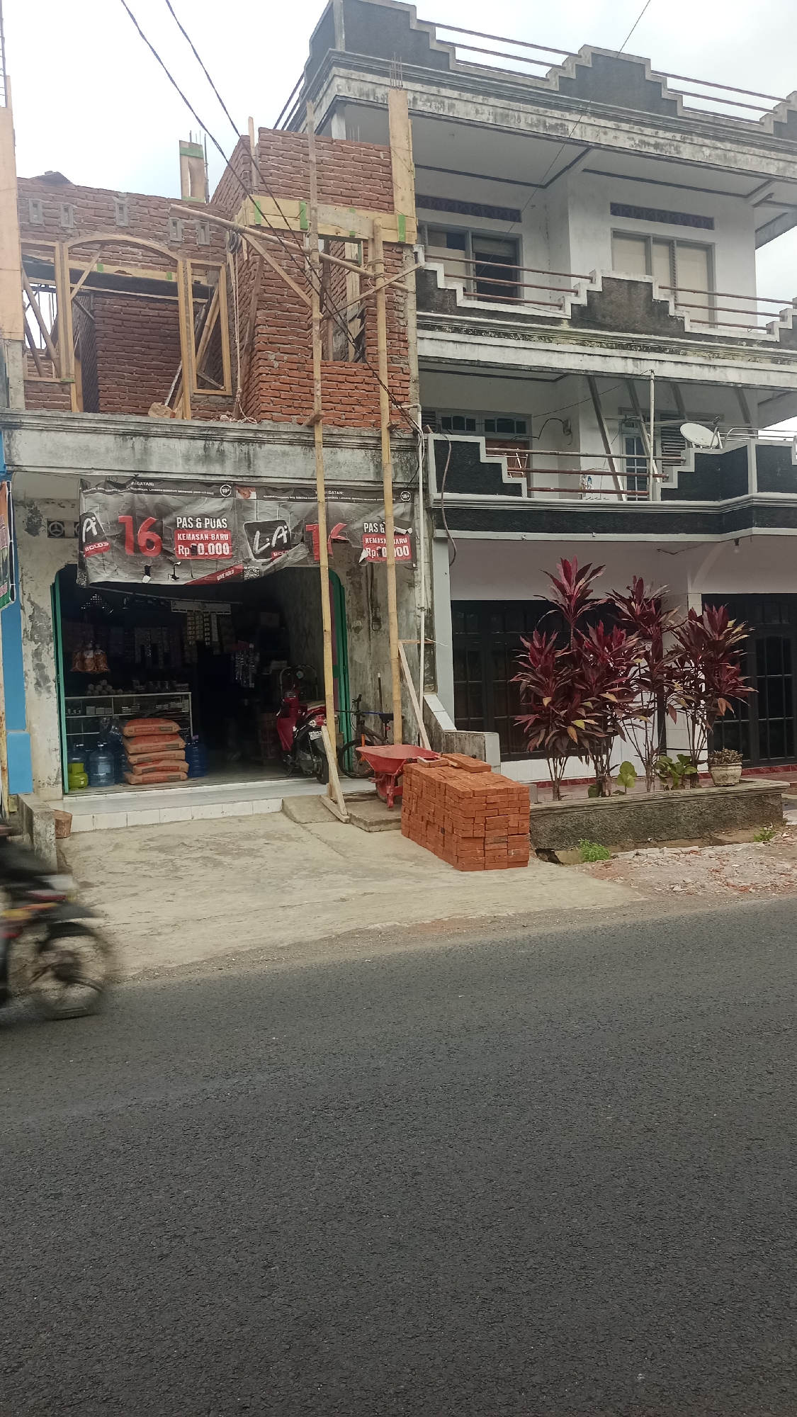 A red motorcycle is blurred by motion on the left side of the frame, suggesting it is moving past a storefront undergoing construction. The storefront, located in Karyamandala, Indonesia, has an open entrance displaying shelves stocked with goods. Bags of cement and water bottles are stacked outside the entrance. Construction materials, including scaffolding and brickwork, are visible on the upper floors of the building. A pile of bricks and a wheelbarrow are positioned on the sidewalk in front of the building. In the background, a multi-story building with balconies and dark-paned windows is partially visible. A planter with dark red foliage stands in front of this building. The scene is set against an overcast sky.