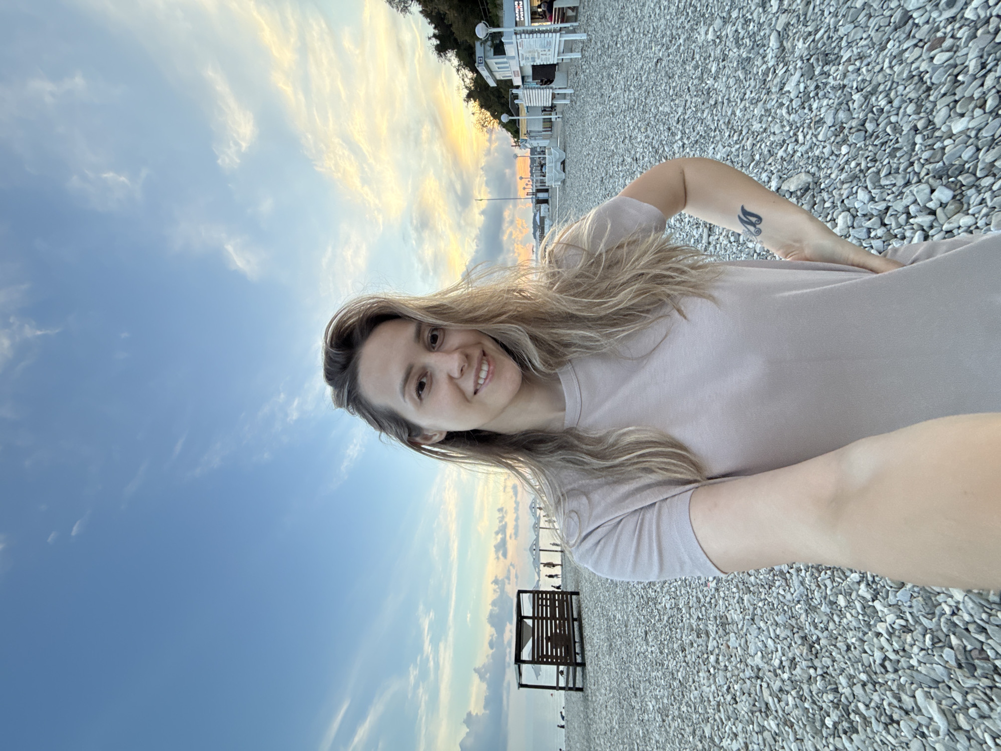 A young Caucasian woman with blonde hair and a light-colored t-shirt is taking a selfie on a pebble beach. She is smiling at the camera, with her left hand on her hip. There is a tattoo on her right forearm. In the background, the sea is visible with a cloudy sky overhead. There are beach umbrellas and some wooden structures on the beach. The location is the city of Nebug, Russia. The overall atmosphere is relaxed and serene.